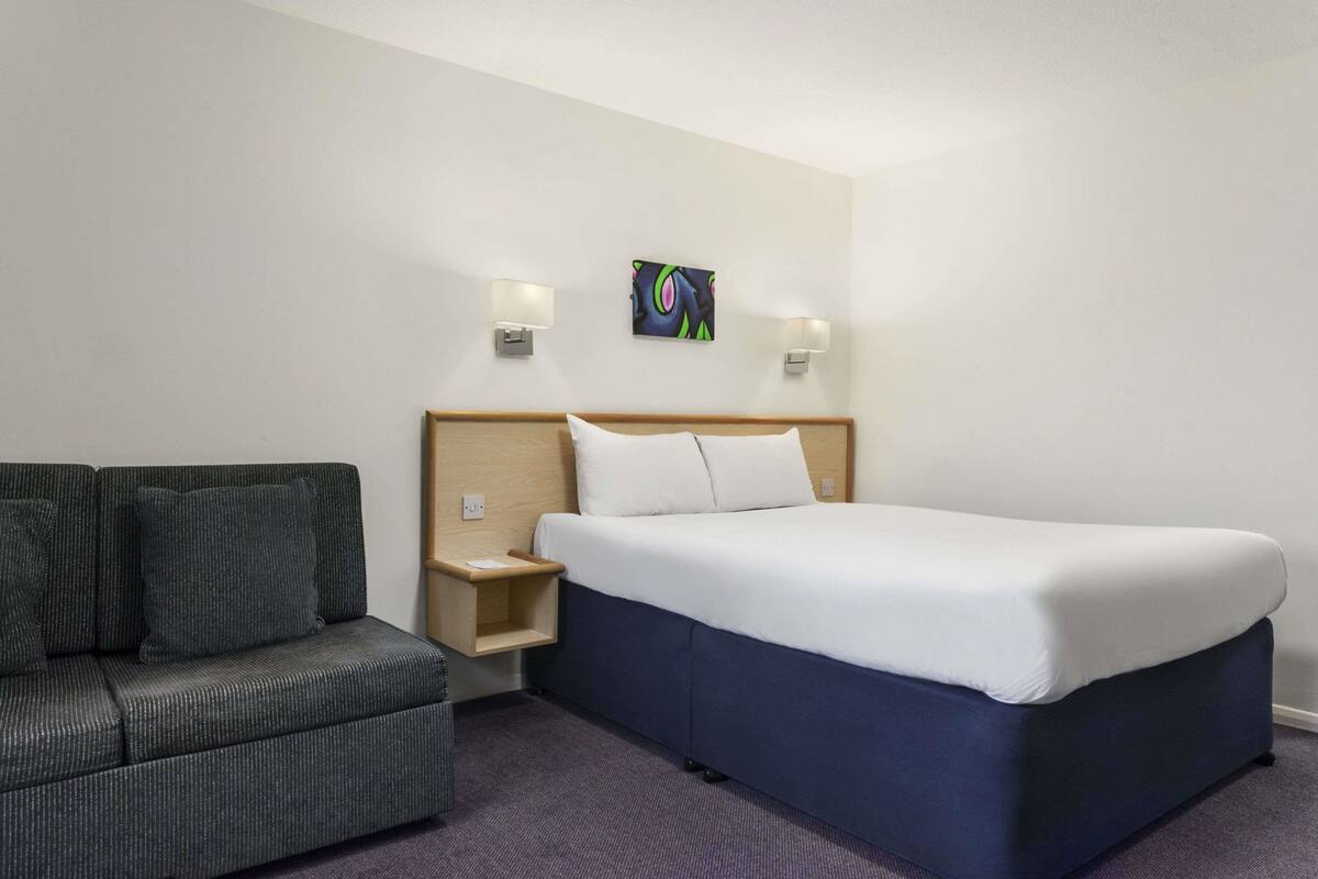 Habitació d'hotel amb llit, sofà, taulella de nit i làmpada penjada a la paret.