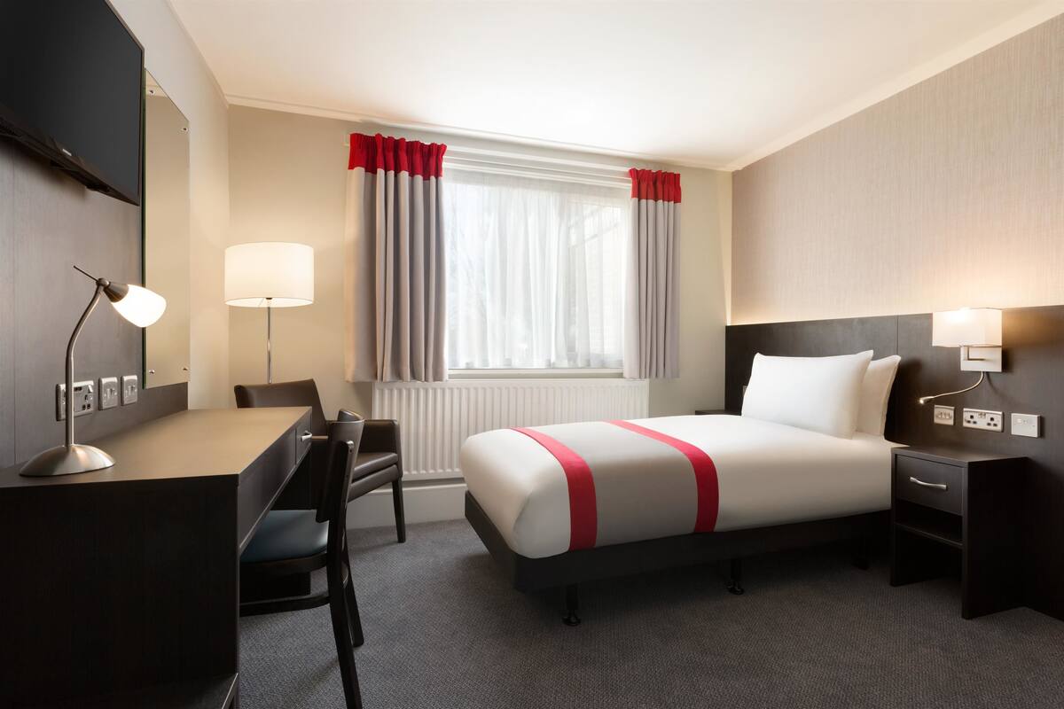 Hotellihuone, jossa on sänky, työpöytä, tuoli, lamppu ja ikkuna, jossa on verhot.