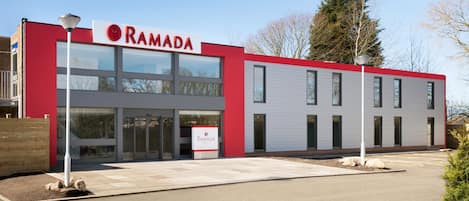 Un edificio moderno con fachada roja y blanca, un letrero que dice "RAMADA" y una zona de entrada pavimentada.