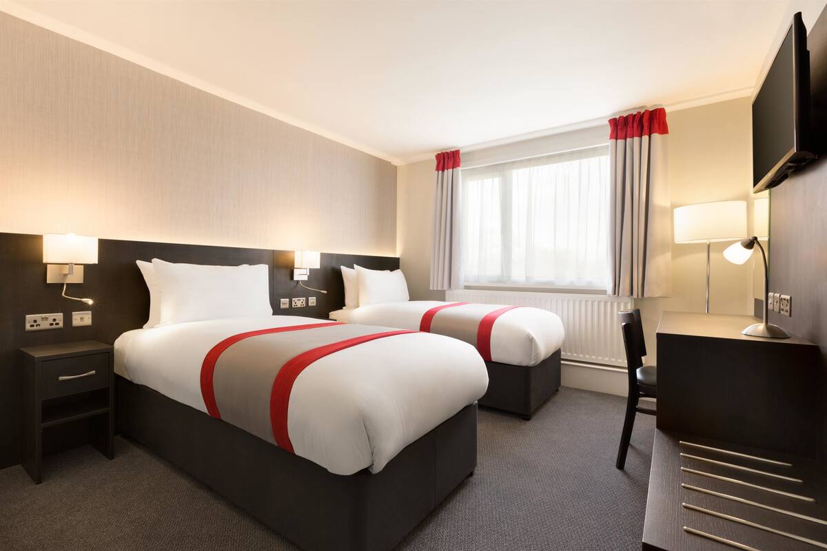 Hotellihuone, jossa on kaksi sänkyä, työpöytä, tuoli, televisio ja ikkuna, jossa on verhot.
