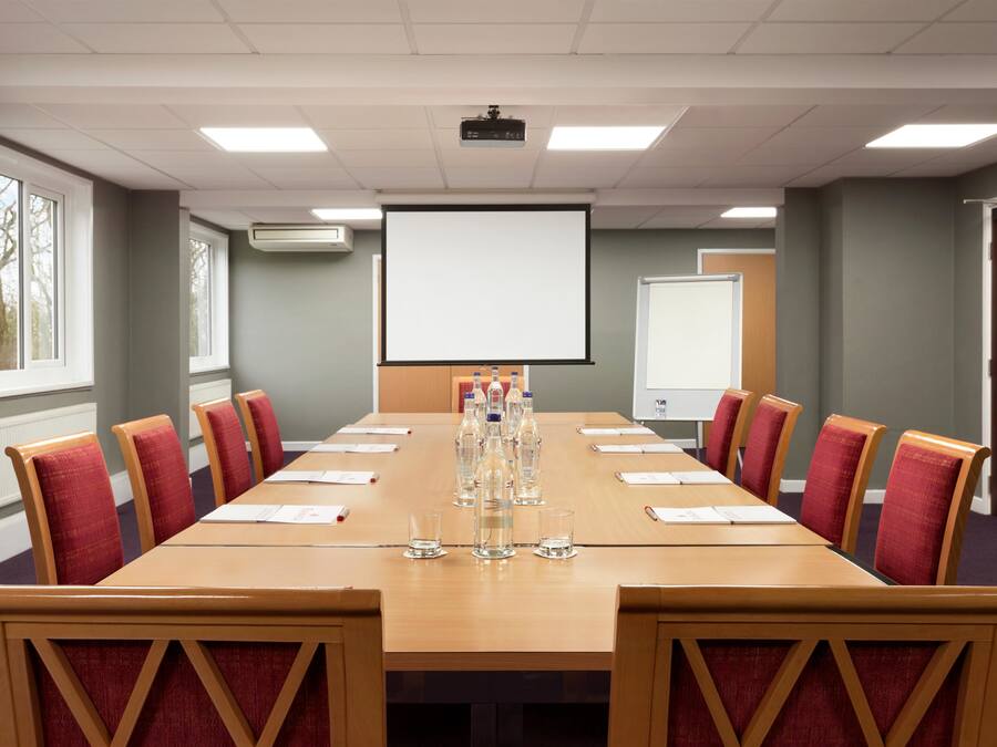 A conference room with a long table, red upholstered chairs, a projector screen, and a whiteboard.