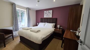 Una cama bien hecha con almohadas y una toalla doblada, una mesita de noche con una lámpara, una silla y una ventana con cortinas.