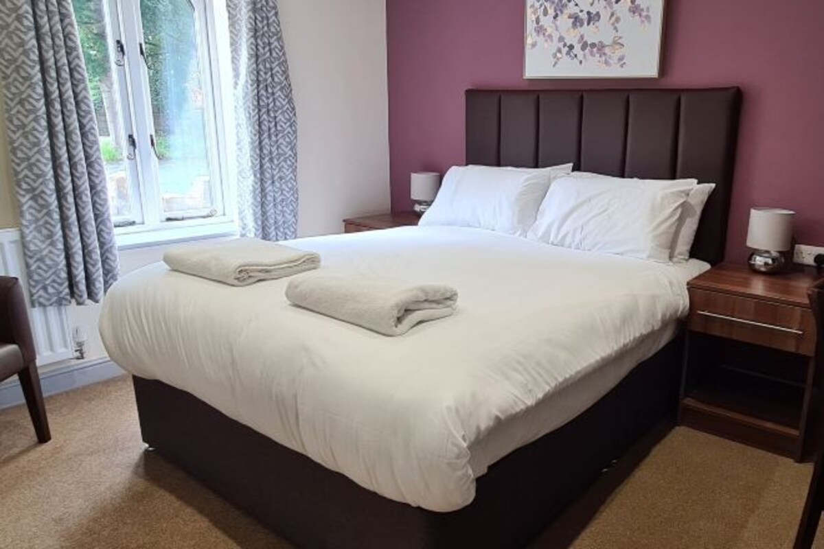Una cama bien tendida con sábanas blancas y una toalla doblada, una mesita de noche con lámpara, y una ventana con cortinas.