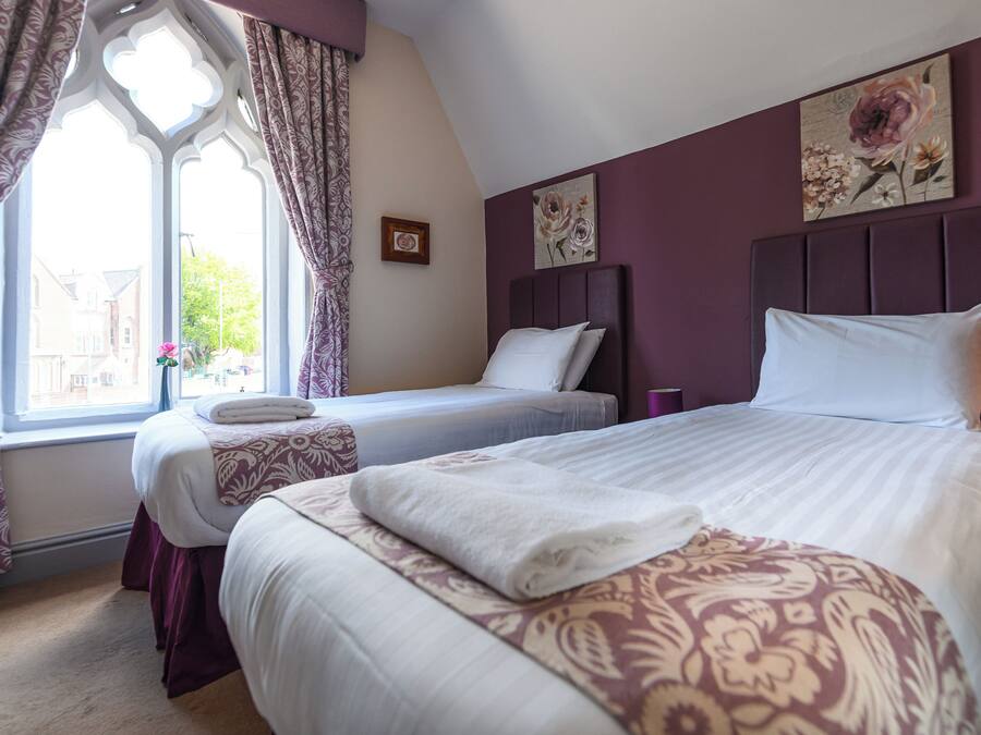 A bedroom with two beds, a window with curtains, and a floral painting on the wall.