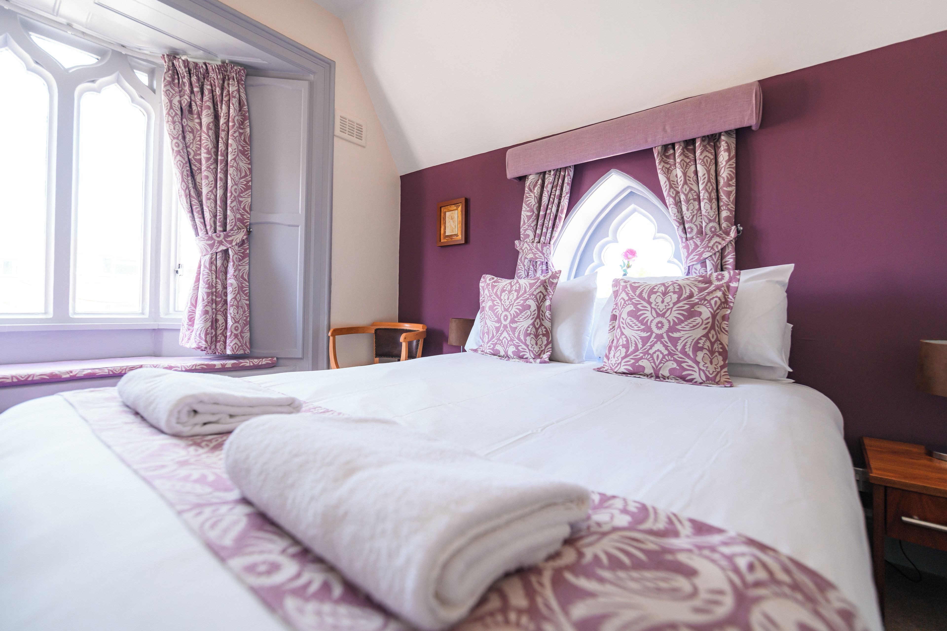 A neatly made bed with patterned bedding, a window with curtains, and a wooden chair.