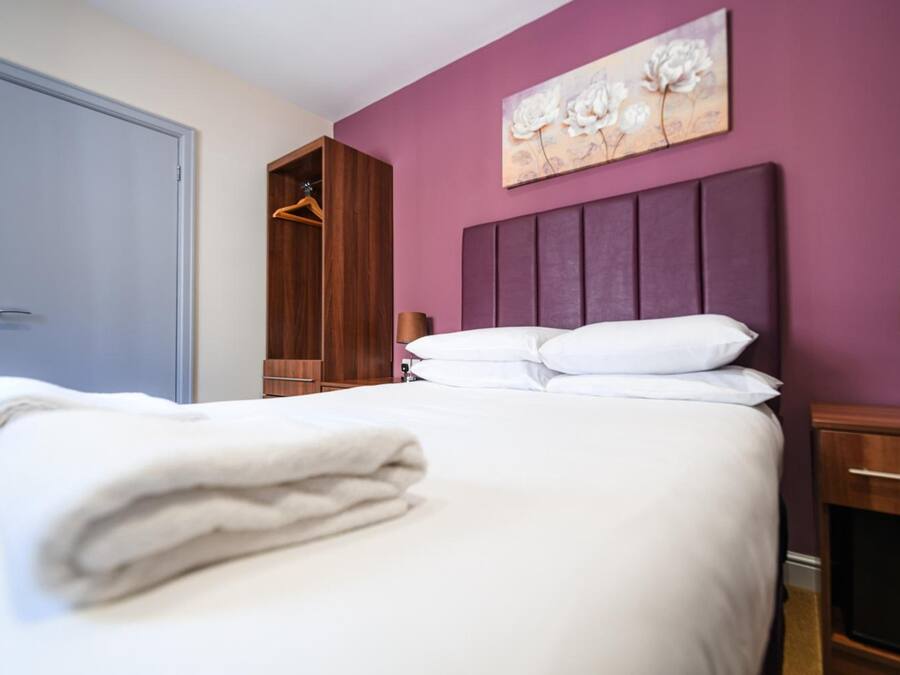 A hotel room with a bed, a wooden wardrobe, a painting on the wall, and a nightstand with a lamp.