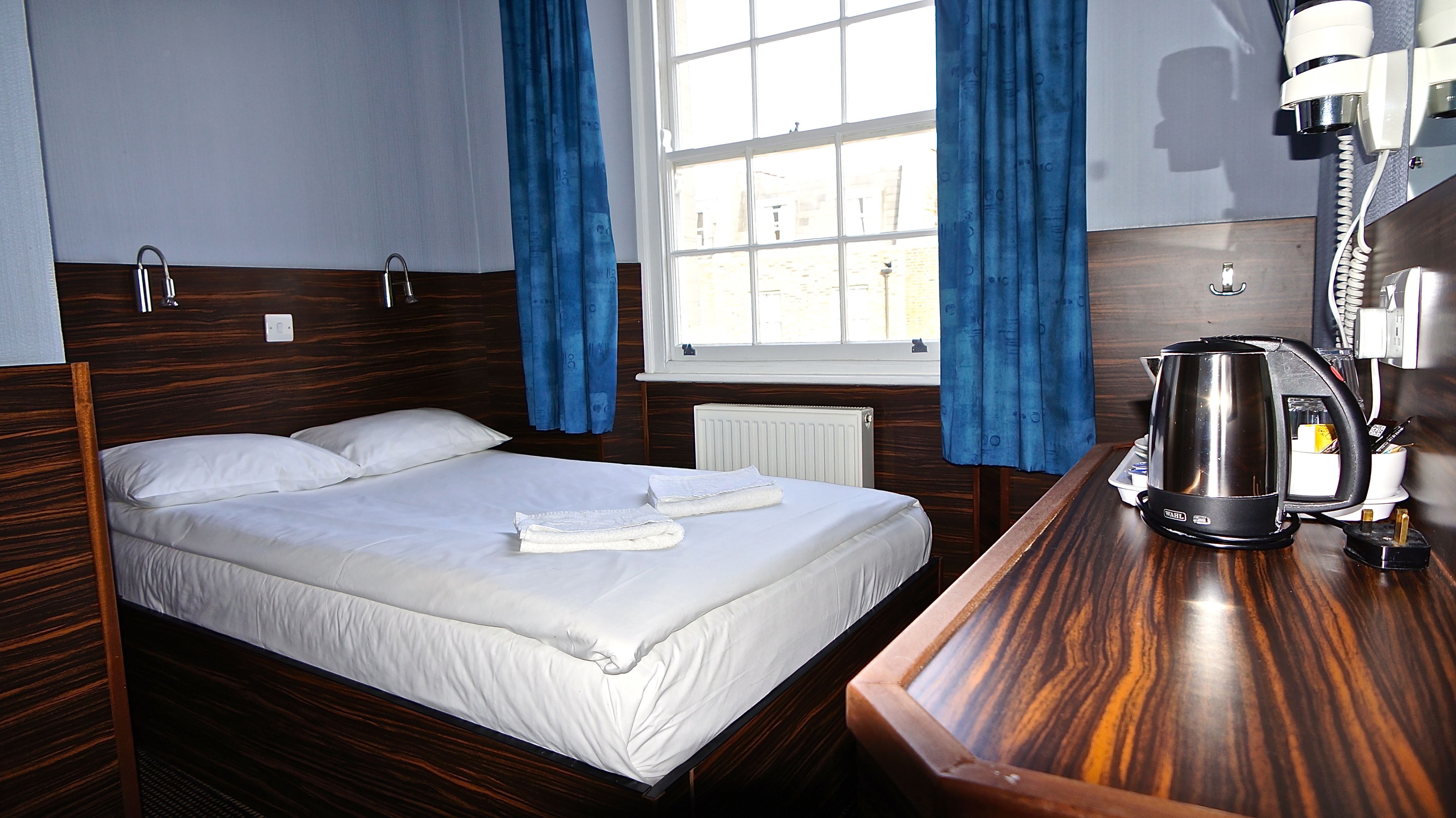 Habitación de hotel con cama de madera, ropa de cama blanca, un radiador, un hervidor de agua y una ventana con cortinas azules.