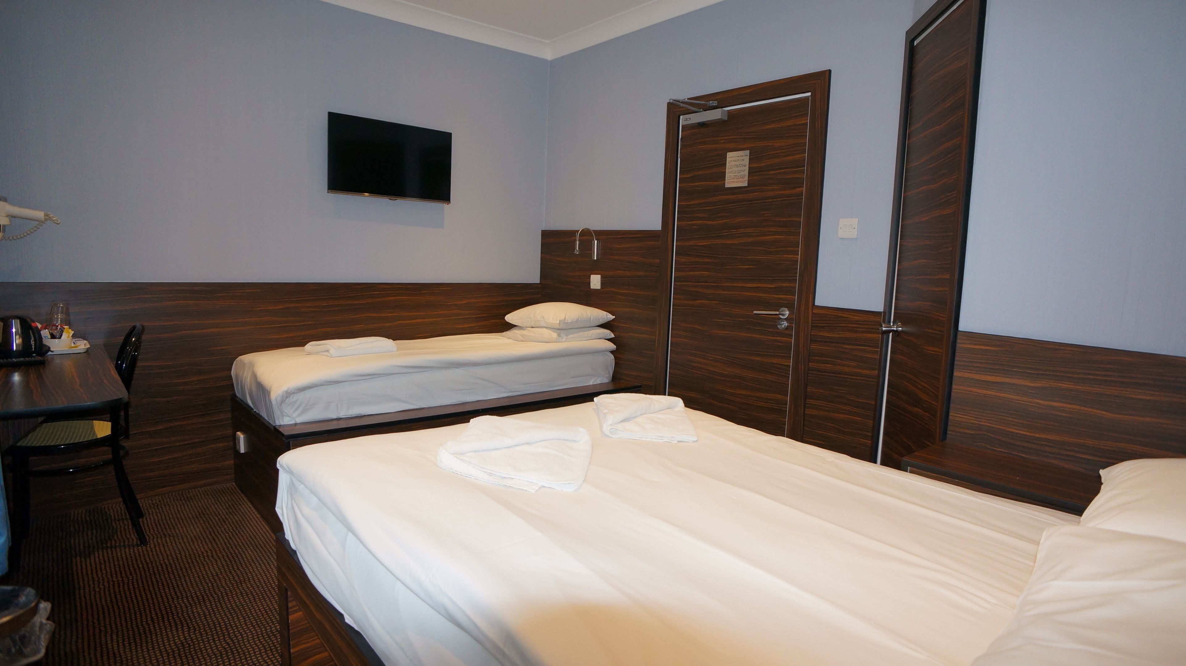 Habitación de hotel con dos camas, un escritorio con silla, un televisor montado en la pared y un armario de madera.