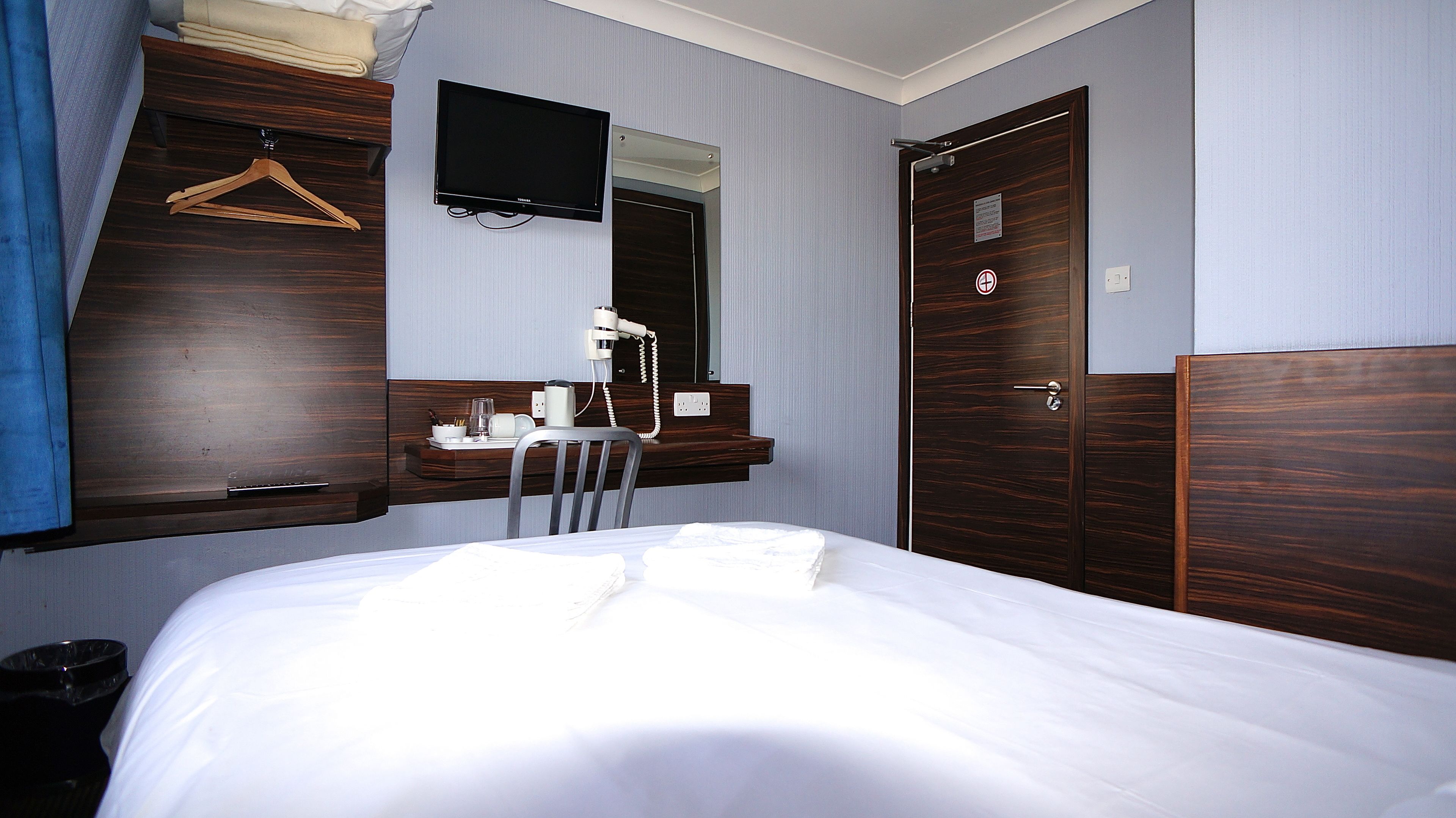 A hotel room with a bed, a desk with a chair, a TV mounted on the wall, and a wardrobe.