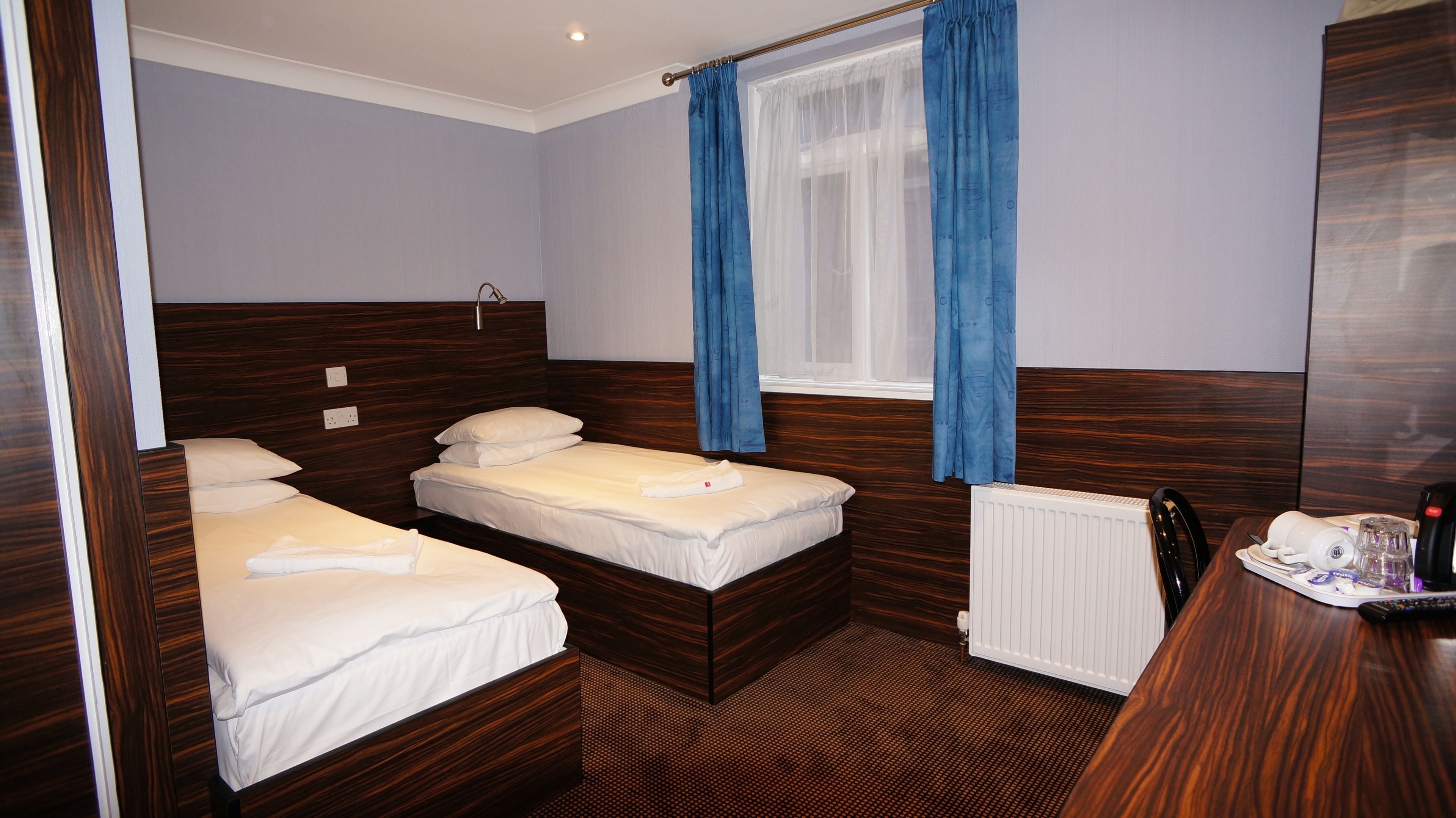 Une chambre d’hôtel avec deux lits simples, une tête de lit en bois, une fenêtre avec des rideaux bleus, un radiateur et une petite table avec une télécommande.