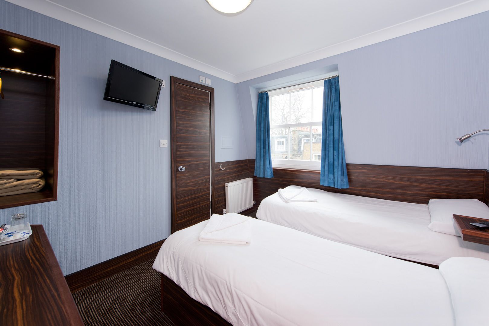 Camera d'albergo con due letti, TV a parete, armadio in legno e finestra con tende blu.