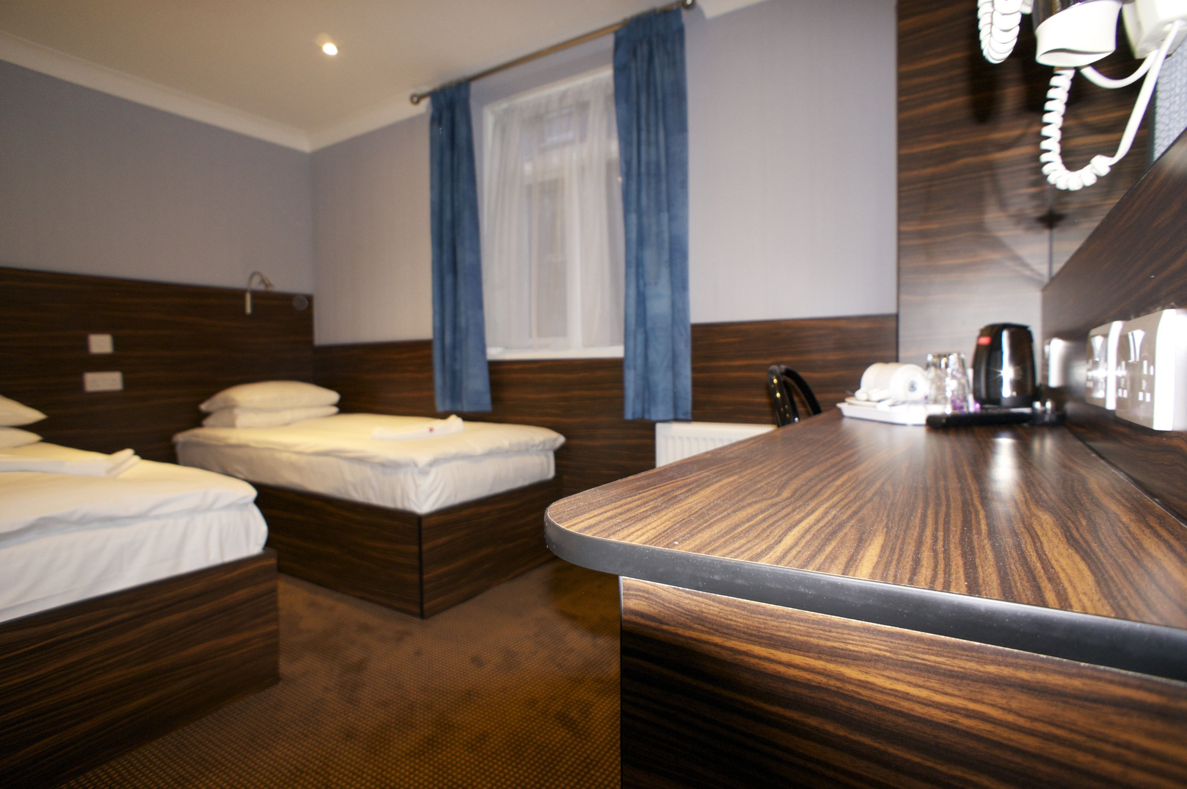 Quarto de hotel com duas camas, uma escrivaninha de madeira, um telefone e uma janela com cortinas.