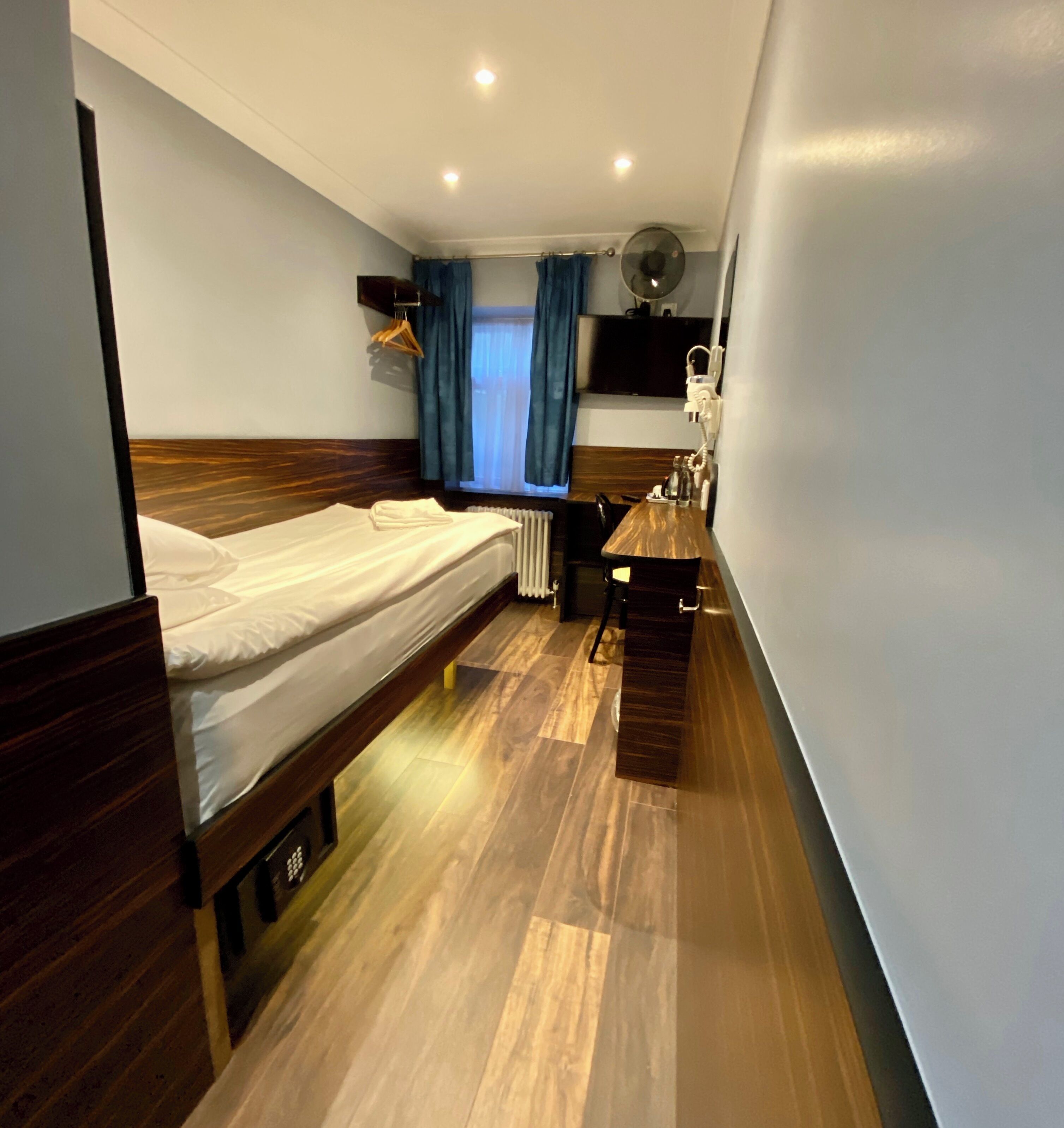 Een compacte hotelkamer met een enkel bed, een klein bureau, een stoel, een nachtkastje, een ventilator en een aan de muur bevestigde televisie.