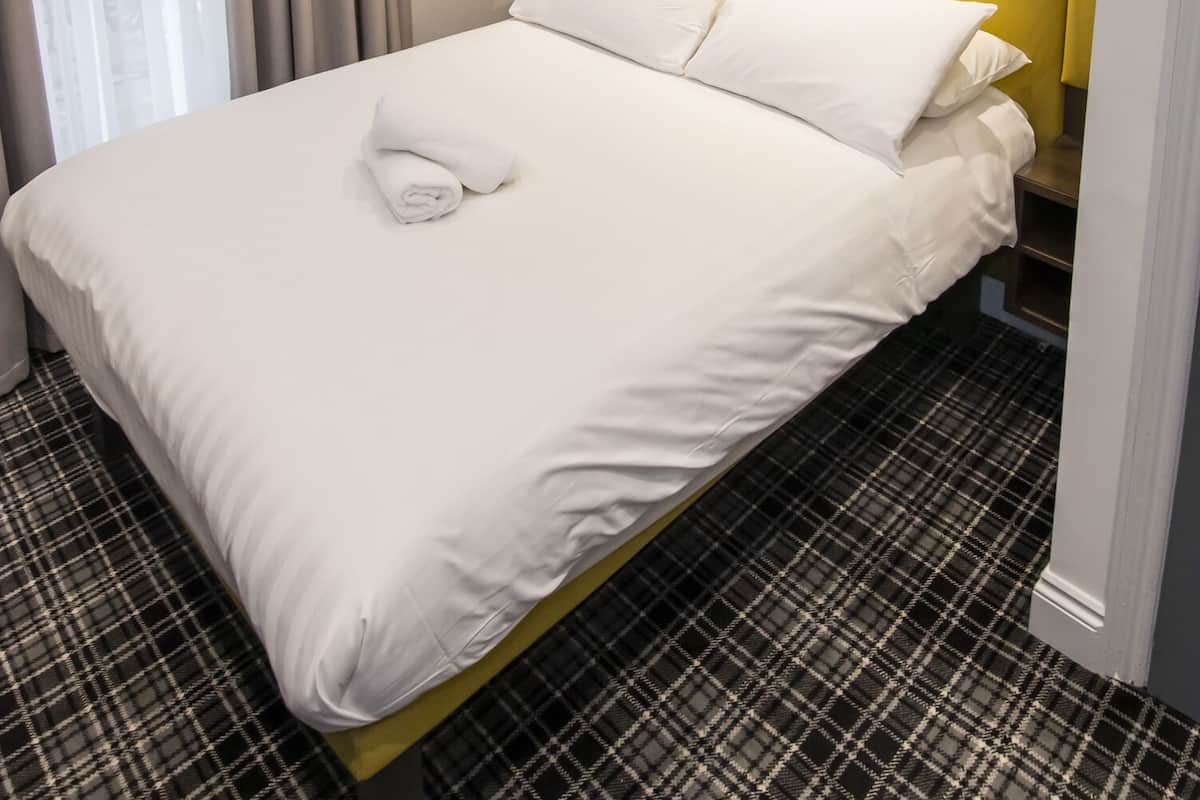 Quarto de hotel moderno com uma cama grande, cabeceira com iluminação integrada, janela com cortinas e piso xadrez.