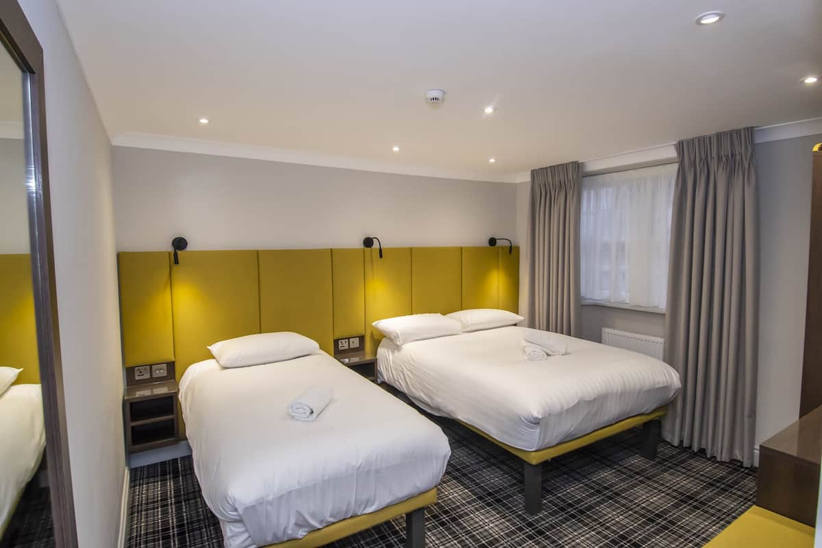 Quarto de hotel com duas camas, cabeceira, abajures, janela com cortinas e um balcão de recepção.