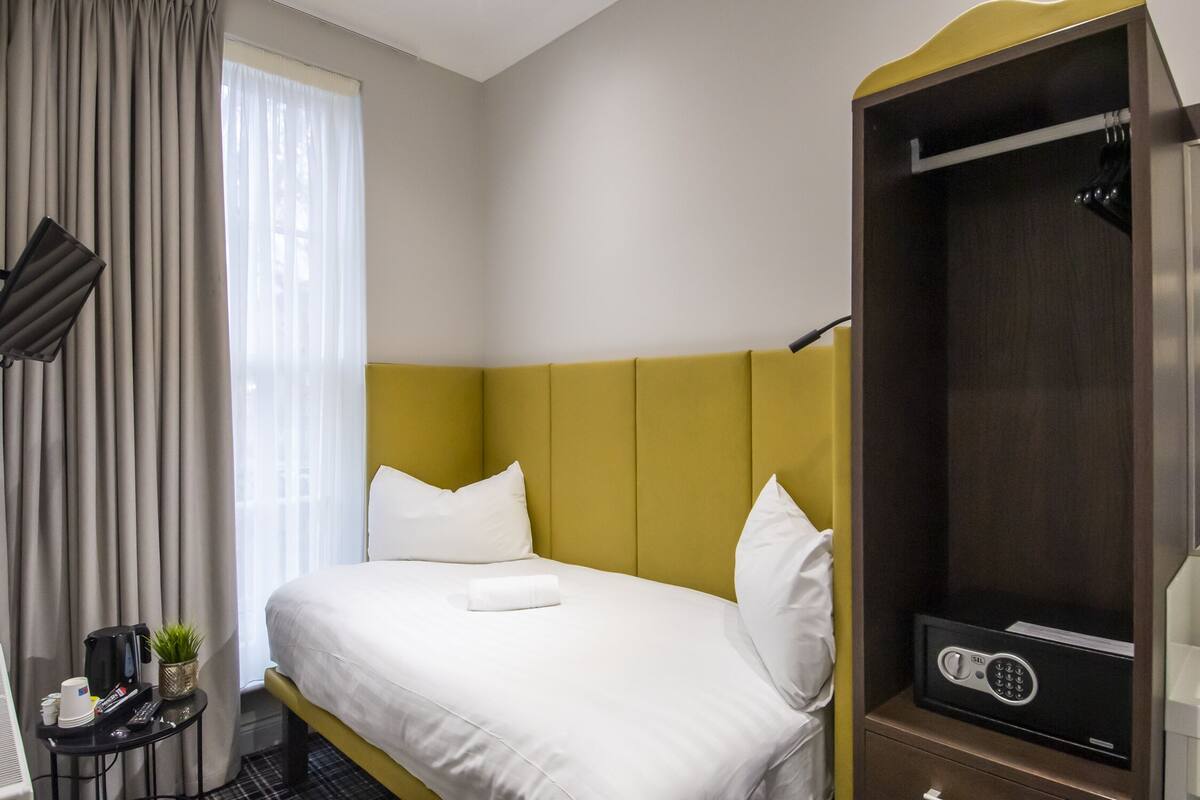 Quarto de hotel com cama, mesa de cabeceira com uma planta, cabideiro e cofre.