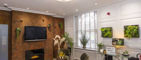 Una sala de estar moderna con chimenea, un televisor grande y una araña. Hay plantas y objetos decorativos en las paredes.