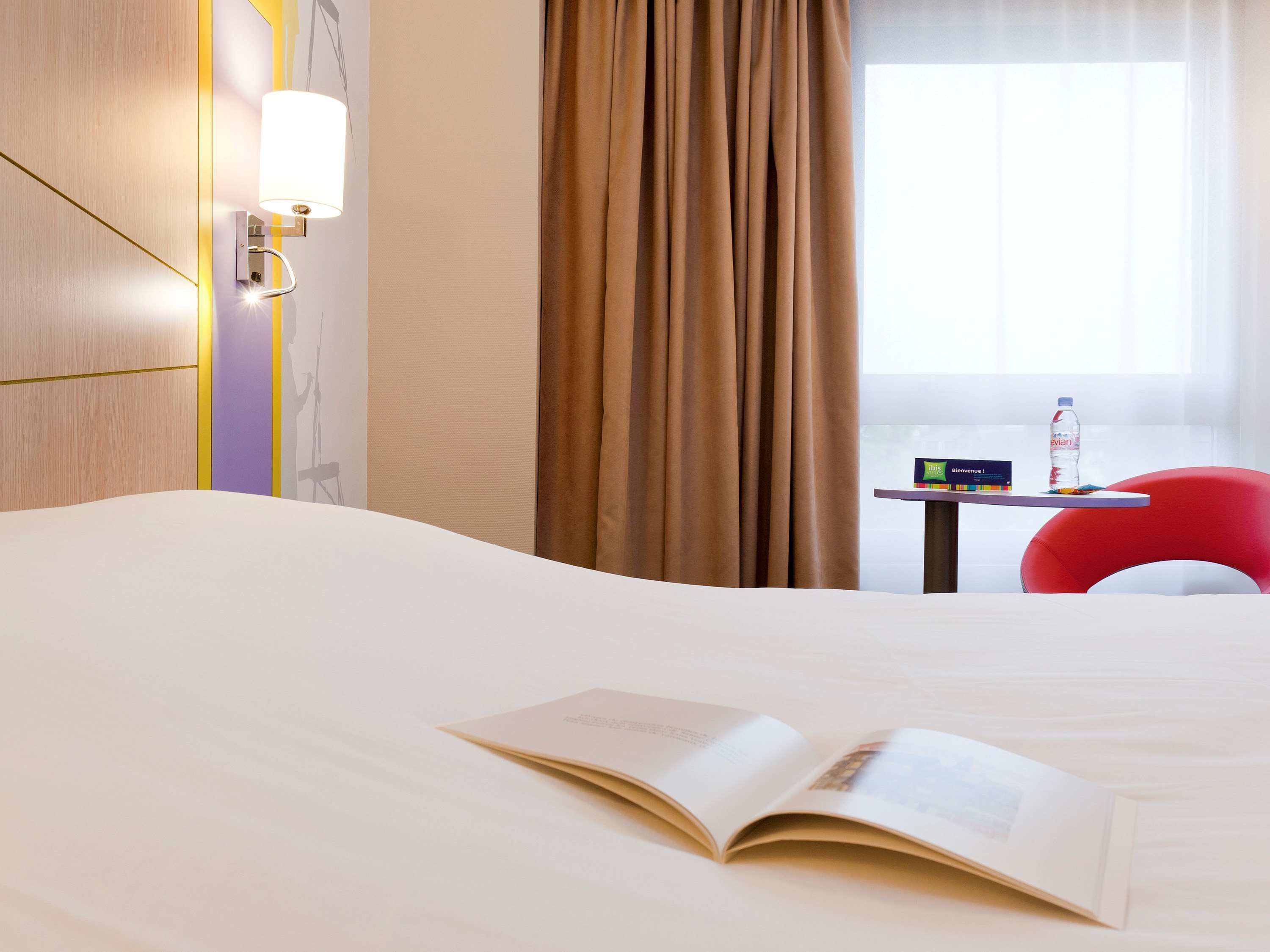 A hotel room with a bed, a table with a bottle of water, a red chair, and a window with curtains.