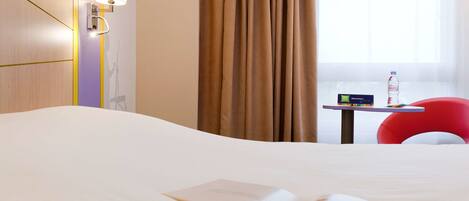 A hotel room with a bed, a table with a bottle of water, a red chair, and a window with curtains.