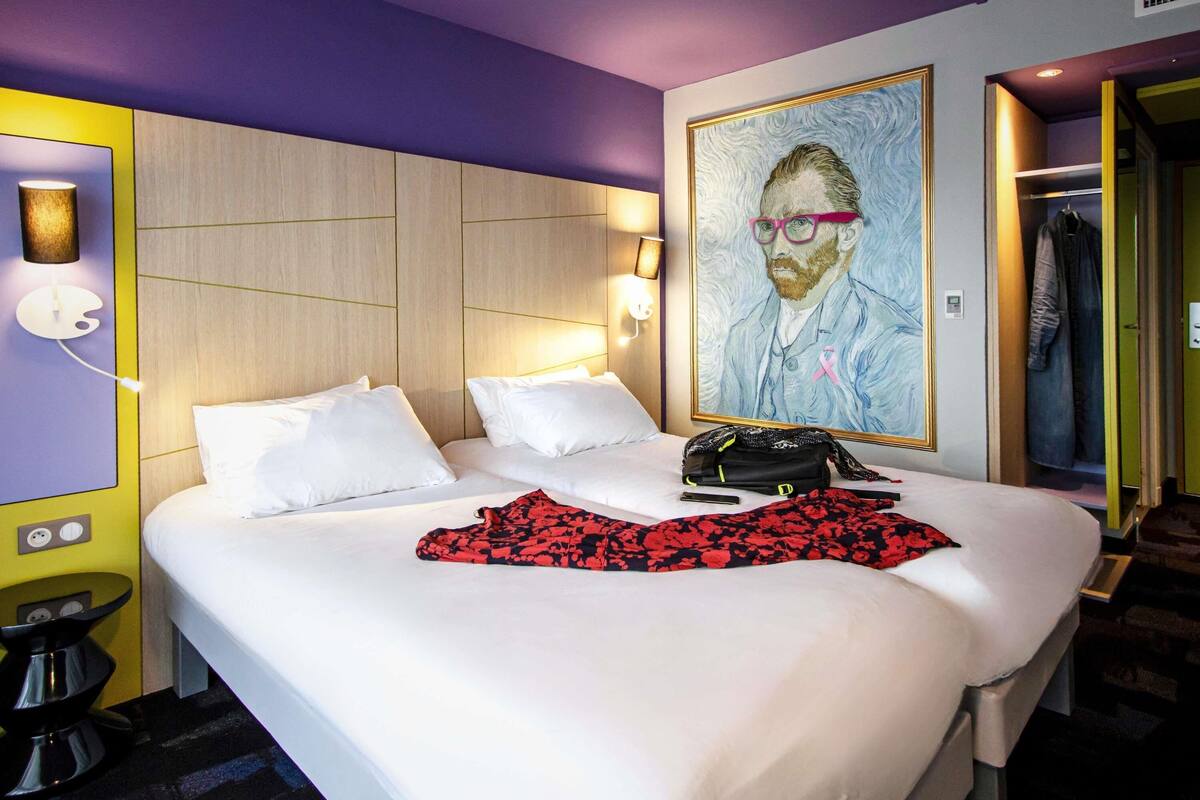 A hotel room with a bed, a painting, a coat rack, and a bathroom door.