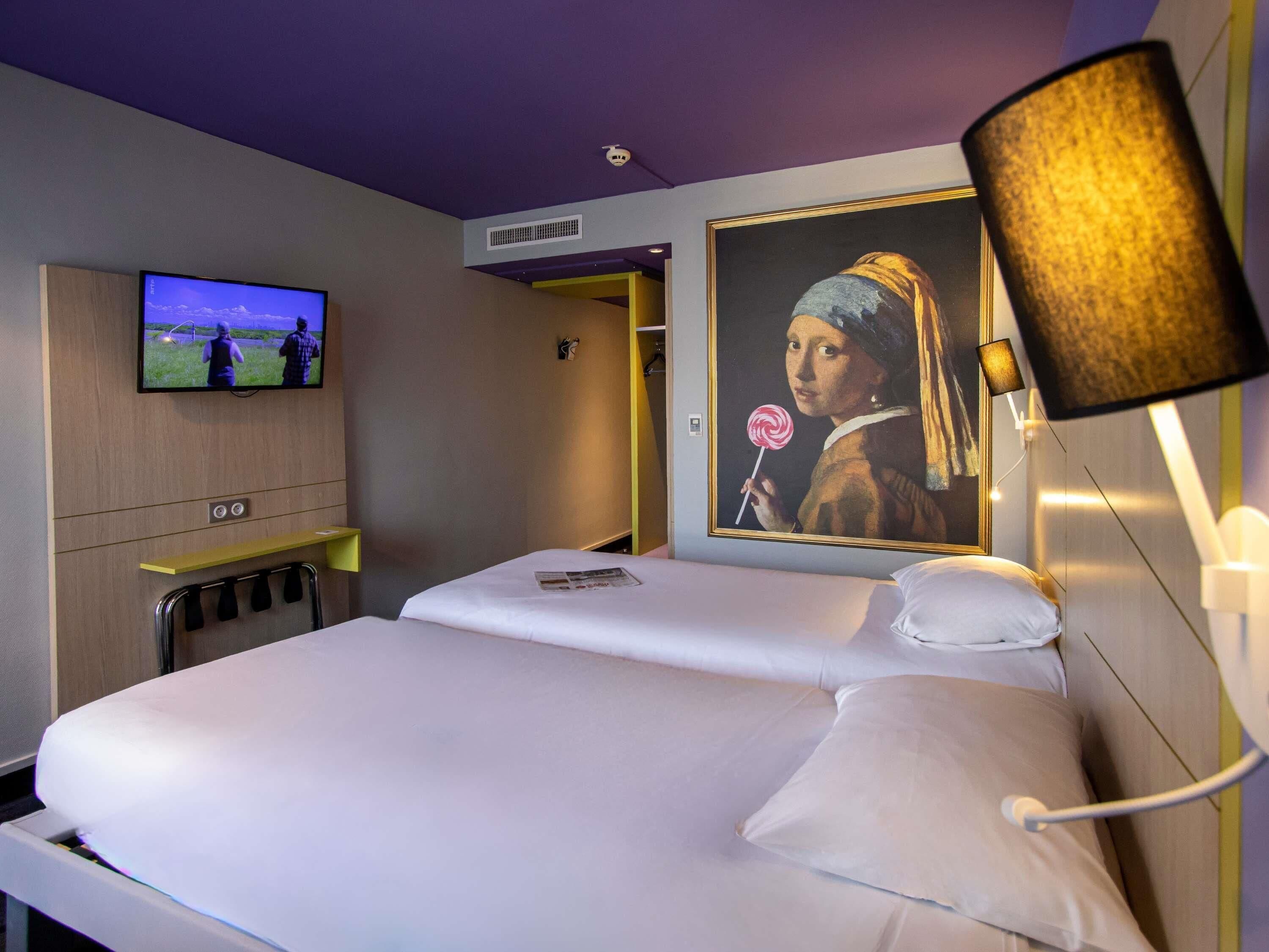 A hotel room with a bed, a television, a painting, and a lamp.