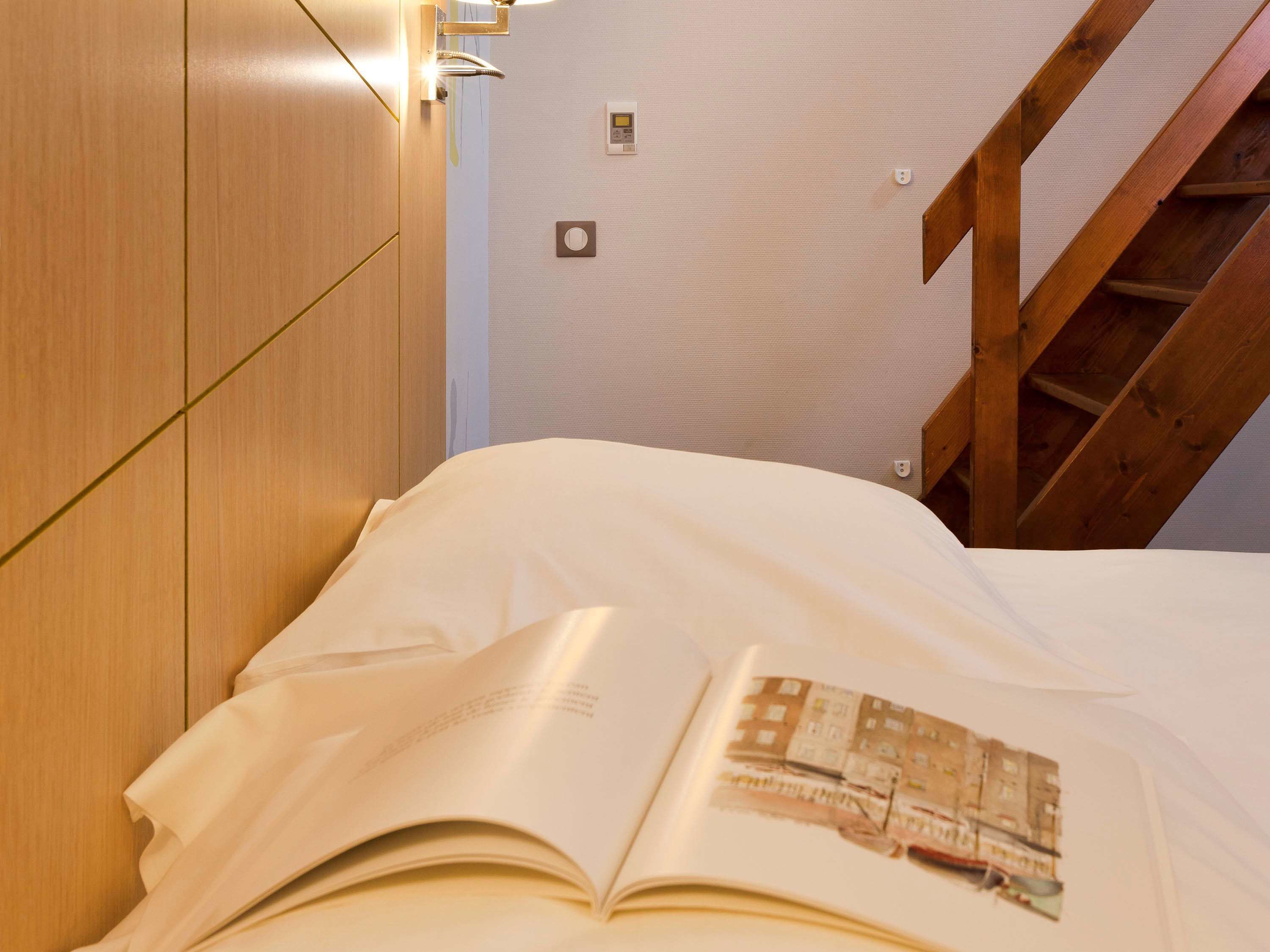 Un lit bien fait, avec un livre ouvert posé dessus, une tête de lit en bois et un escalier en arrière-plan.