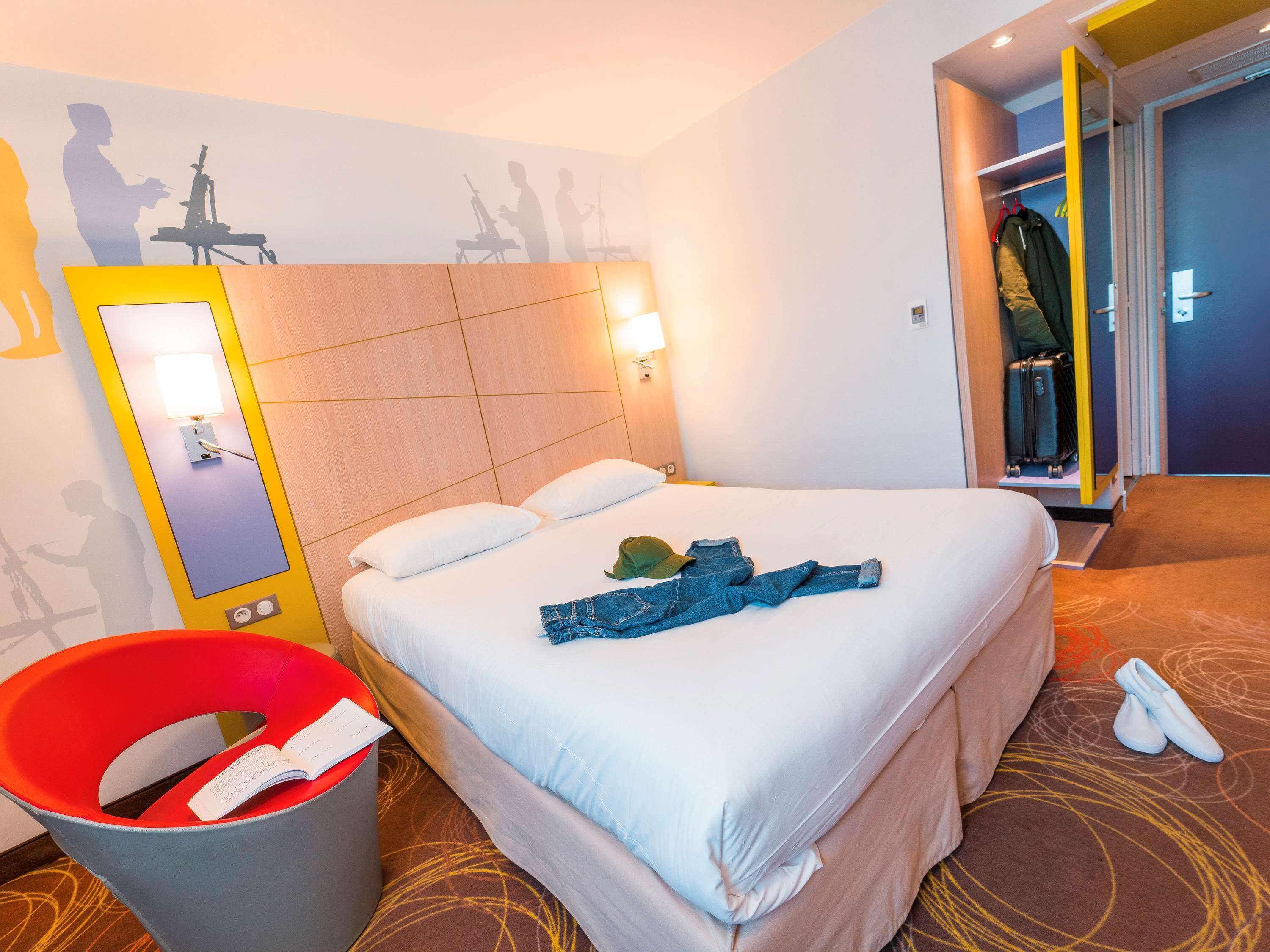 Habitación de hotel con cama, espejo, lámpara, basurero y perchero con chaqueta y maleta.