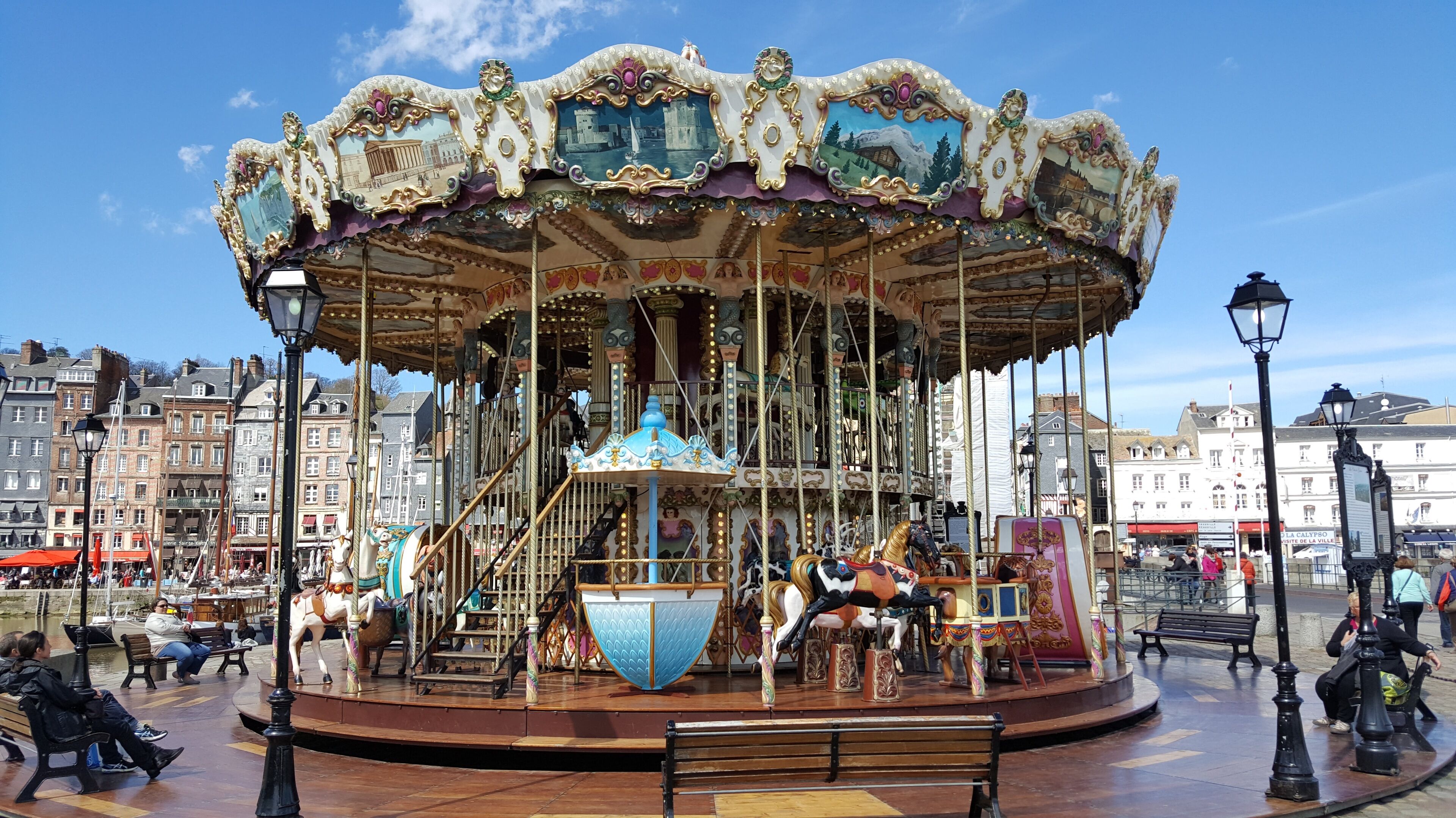 A carousel with ornate decorations and horses, surrounded by benches and a waterfront view.