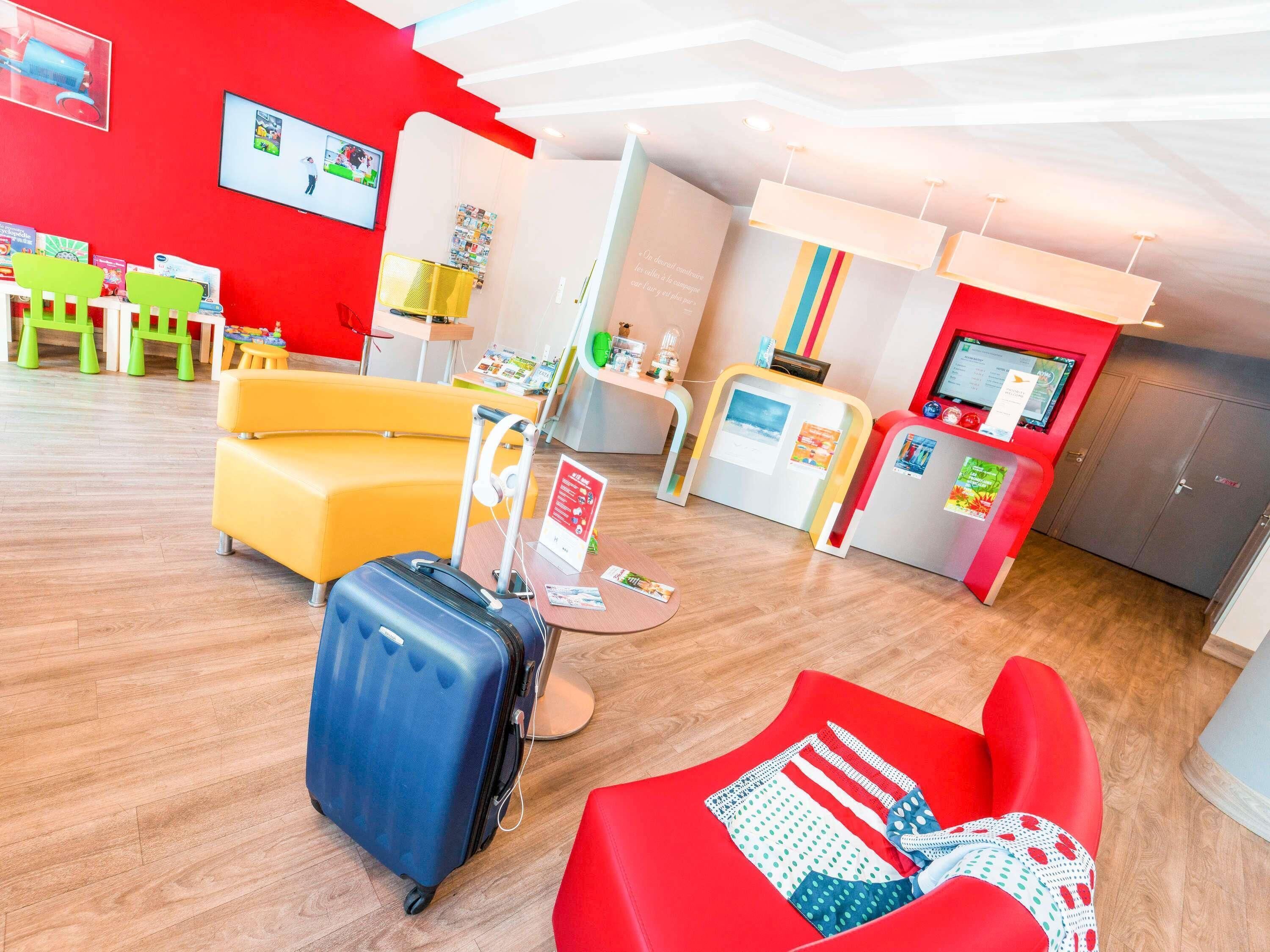 A hotel lobby with a red wall, a yellow sofa, a red chair, a blue suitcase, and a reception desk with a computer monitor.