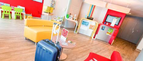 A hotel lobby with a red wall, a yellow sofa, a red chair, a blue suitcase, and a reception desk with a computer monitor.