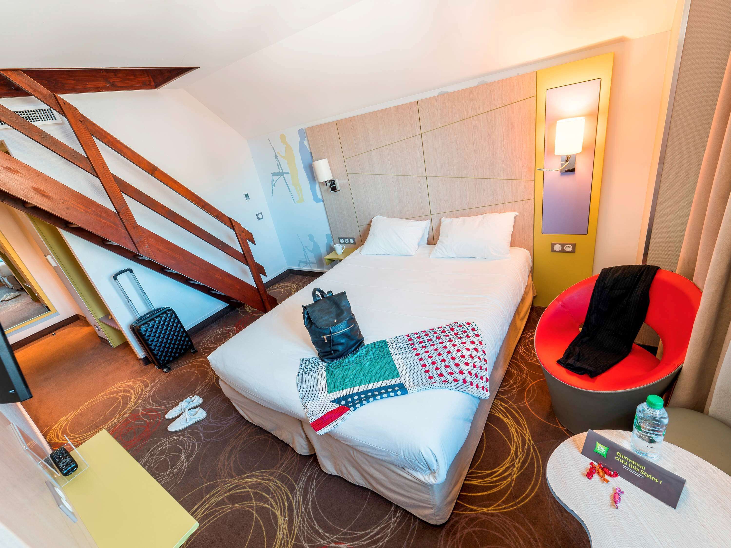 A hotel room with a bed, a chair, a desk, and a small table with a water bottle and a welcome card.