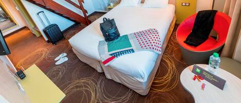 A hotel room with a bed, a chair, a desk, and a small table with a water bottle and a welcome card.