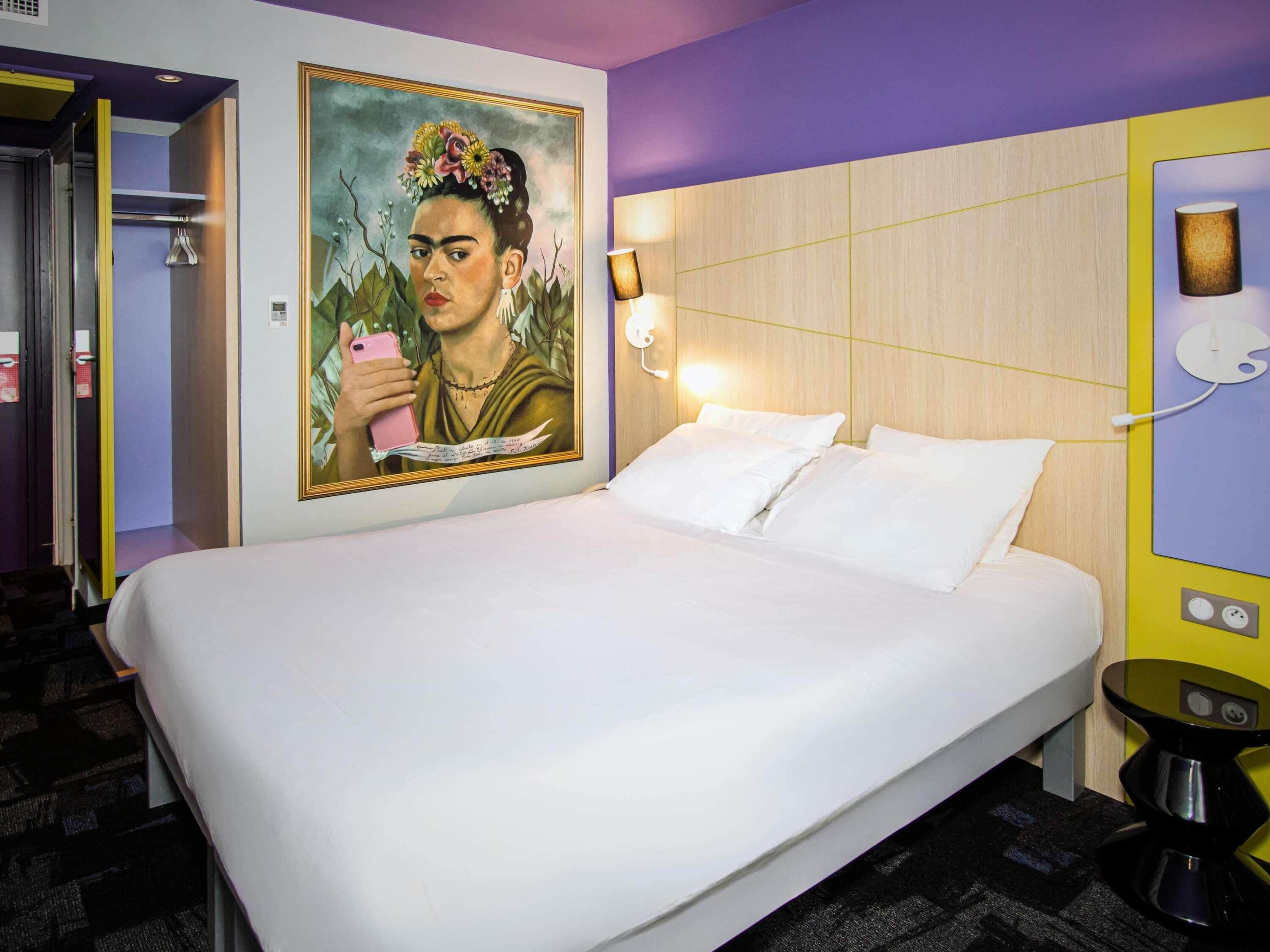 A hotel room with a large bed, a wall-mounted painting, a bedside lamp, and a small table with a lamp.