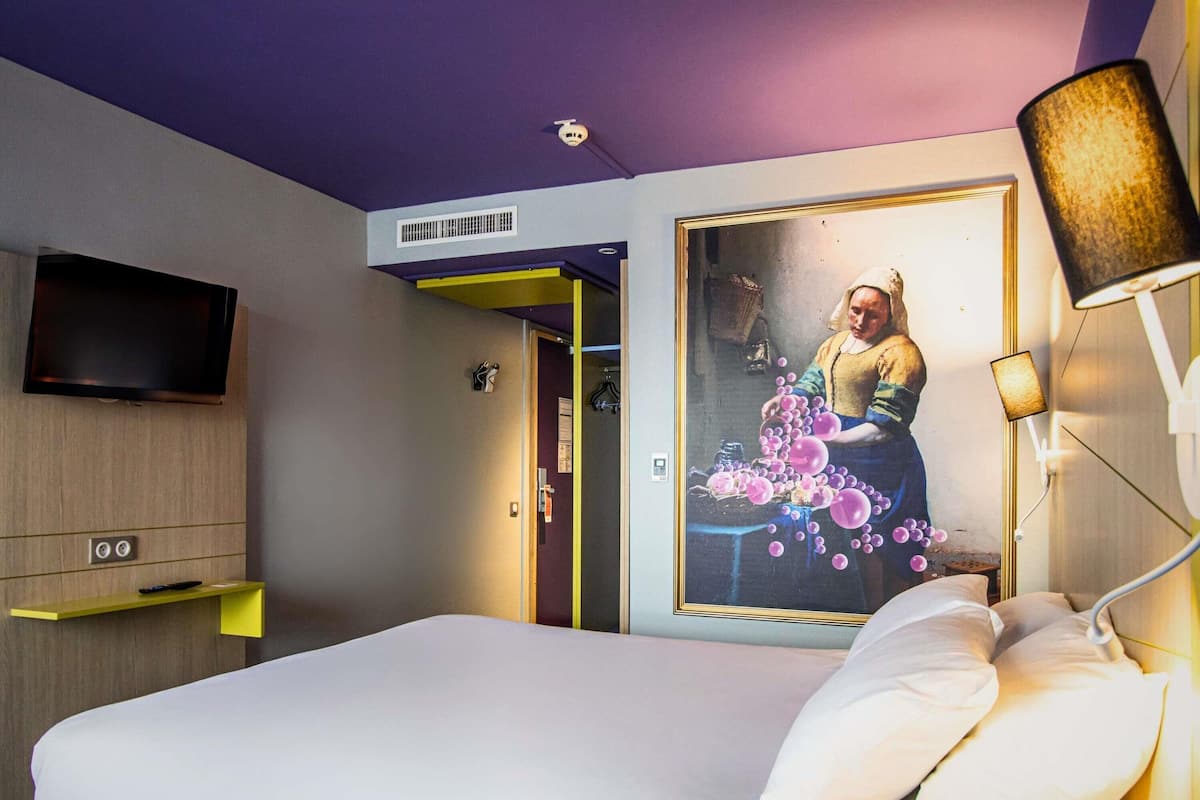 A modern hotel room with a large bed, a flat-screen TV mounted on the wall, and a unique painting of a woman with bubbles.