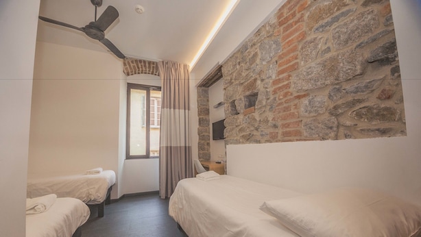Habitación de hotel con dos camas individuales, ventilador de techo, pared de piedra, televisor y ventana con cortinas.