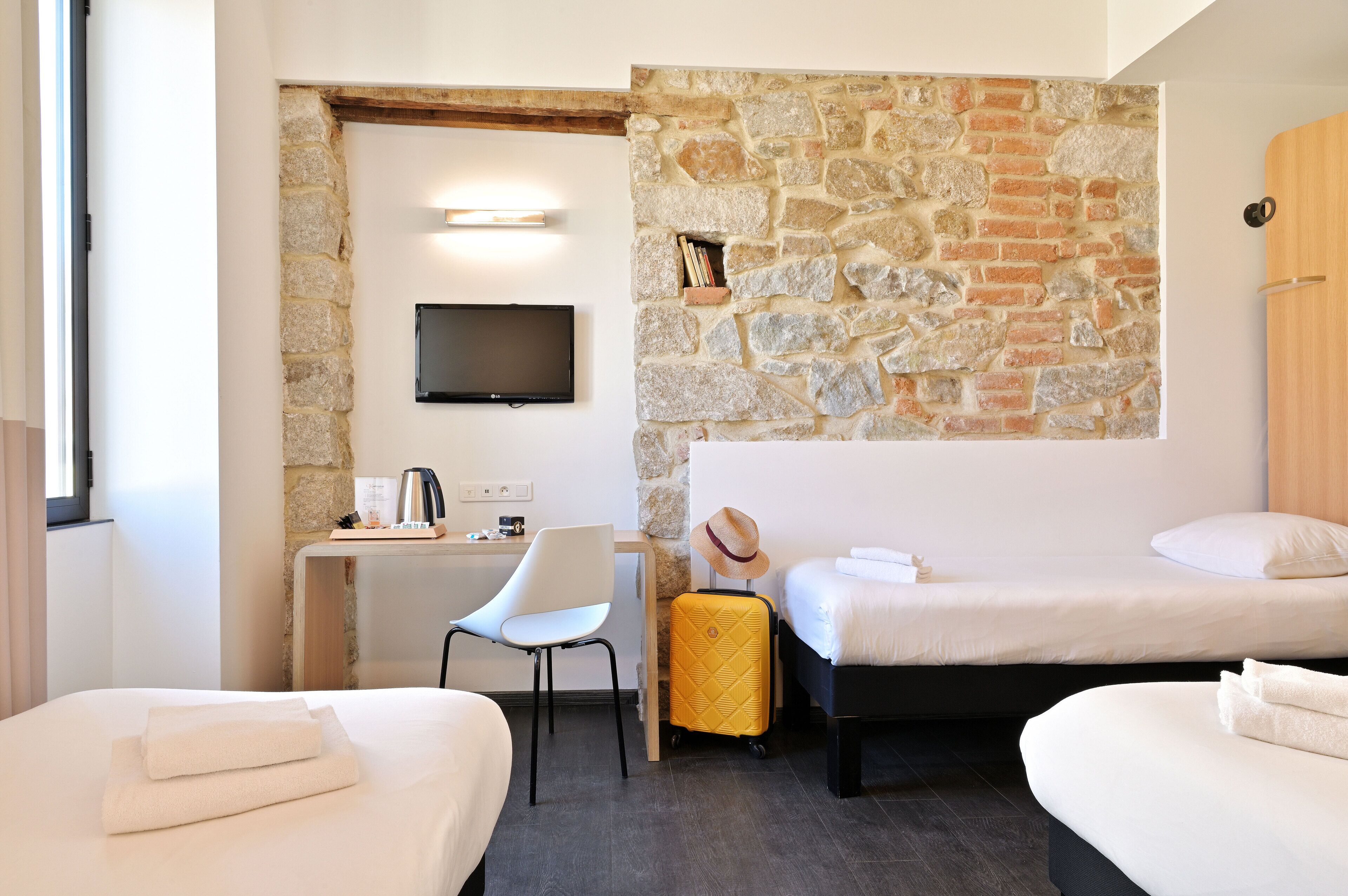 A hotel room with two beds, a desk, a chair, a television, and a stone wall.
