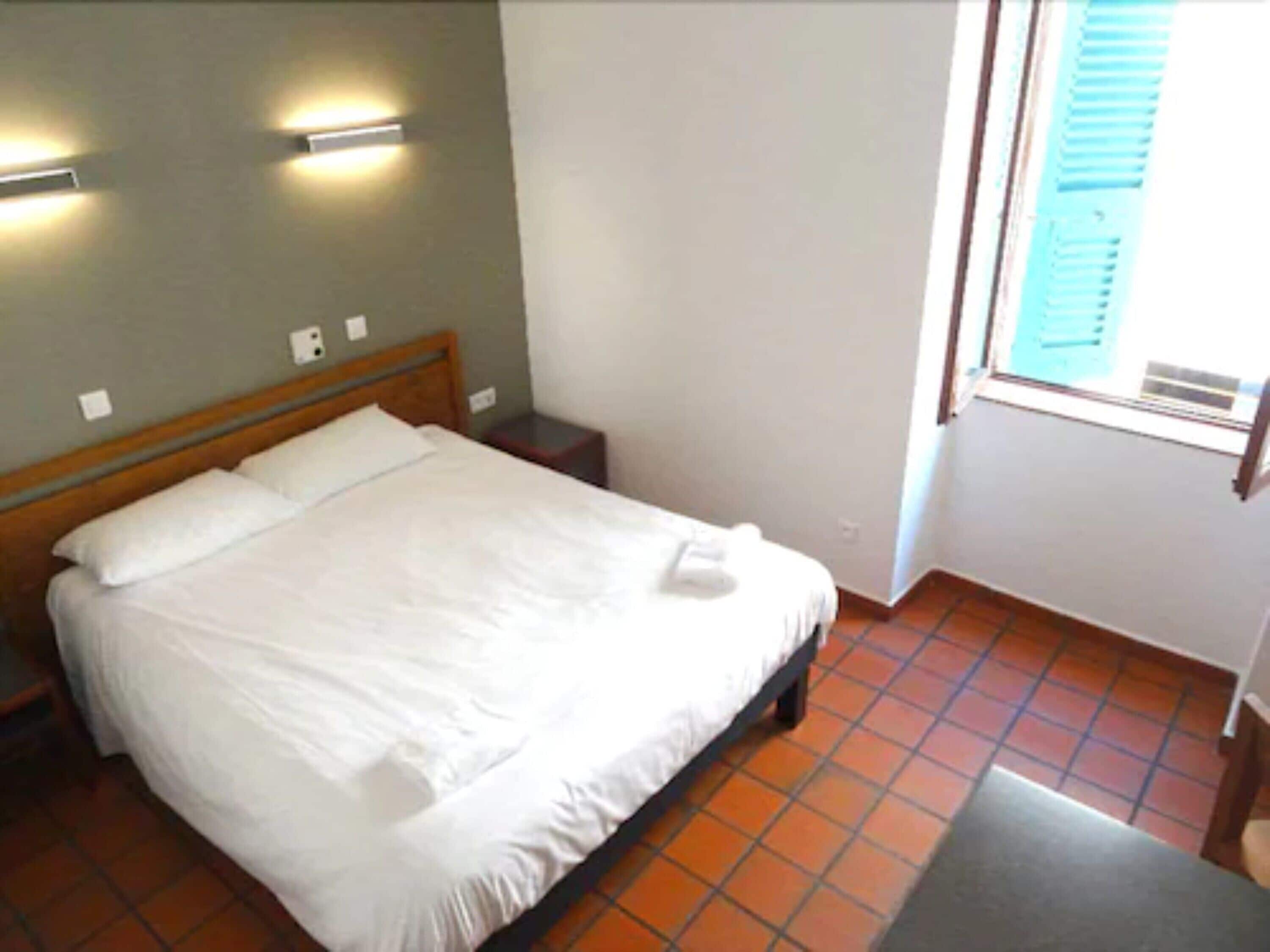 A neatly made bed in a room with a tiled floor and a window with blinds.