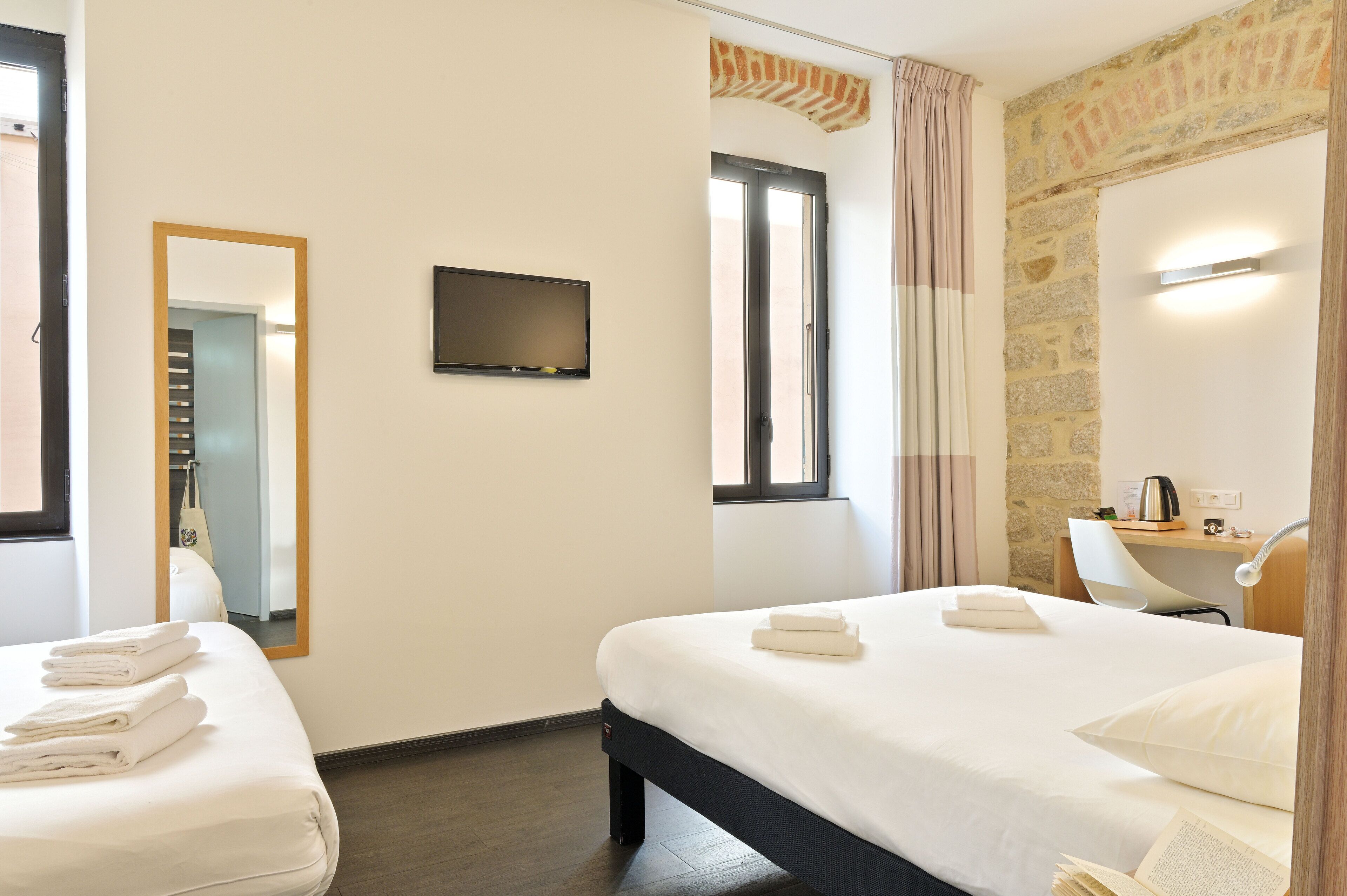 A hotel room with two beds, a flat-screen TV, a desk with a chair, a mirror, and a stone wall.