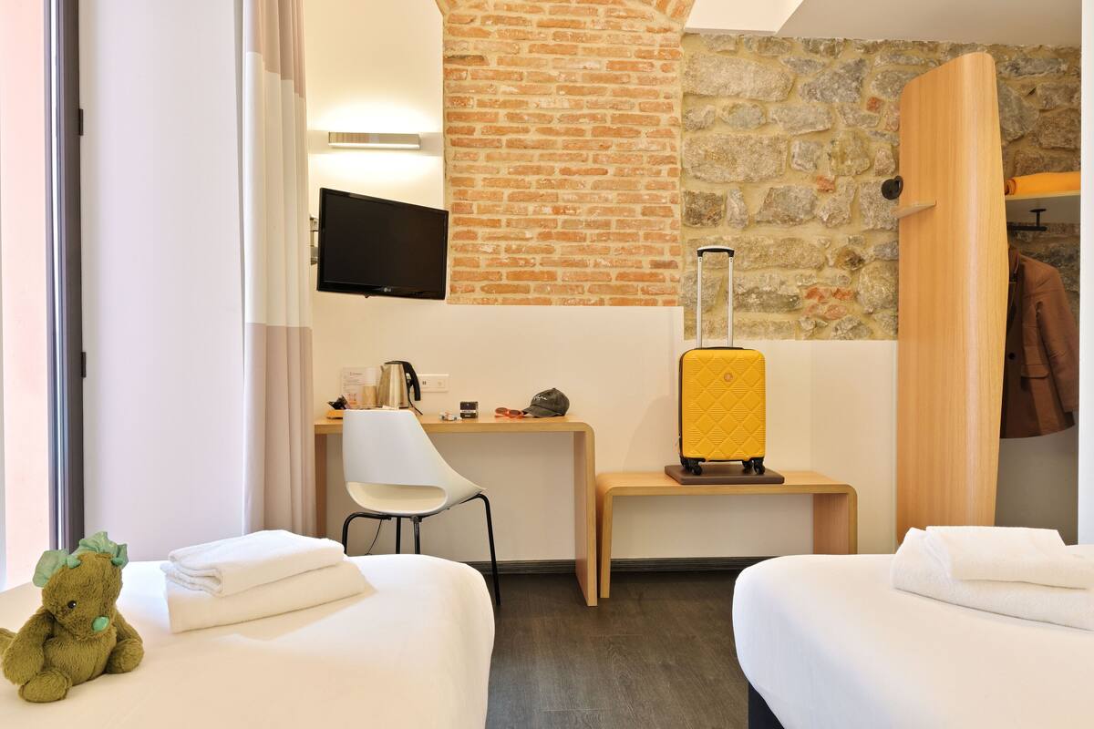 A hotel room with a brick wall, a desk with a chair, a bed, a suitcase, and a teddy bear.