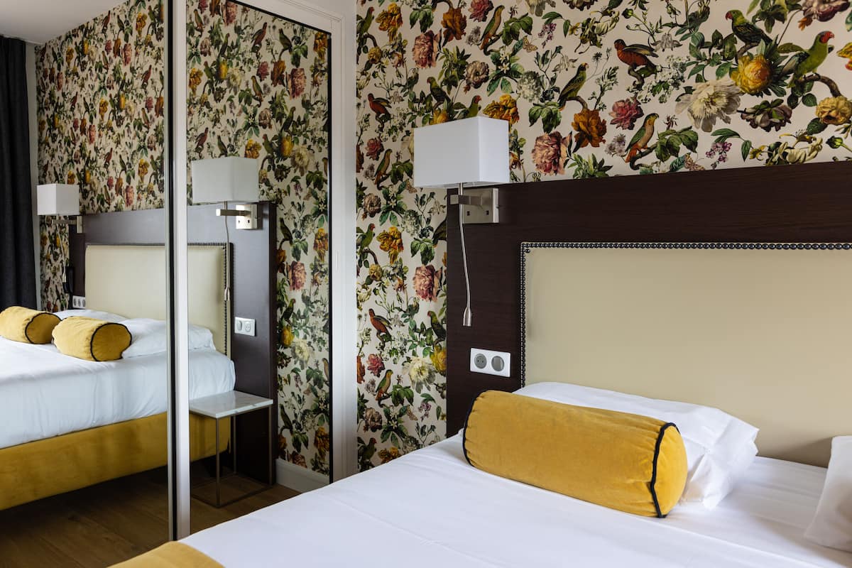 Ein Hotelzimmer mit floralem Wandpapier, einem Bett mit weißen Leinentüchern und einem gelben Zierkissen, einem Nachttisch mit Lampe und einem Spiegel an der Wand.