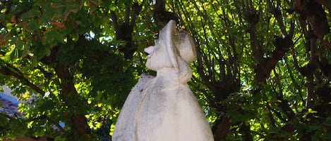 A stone statue of a woman with a book, set against a backdrop of a tree with green leaves and a stone building with a sign.