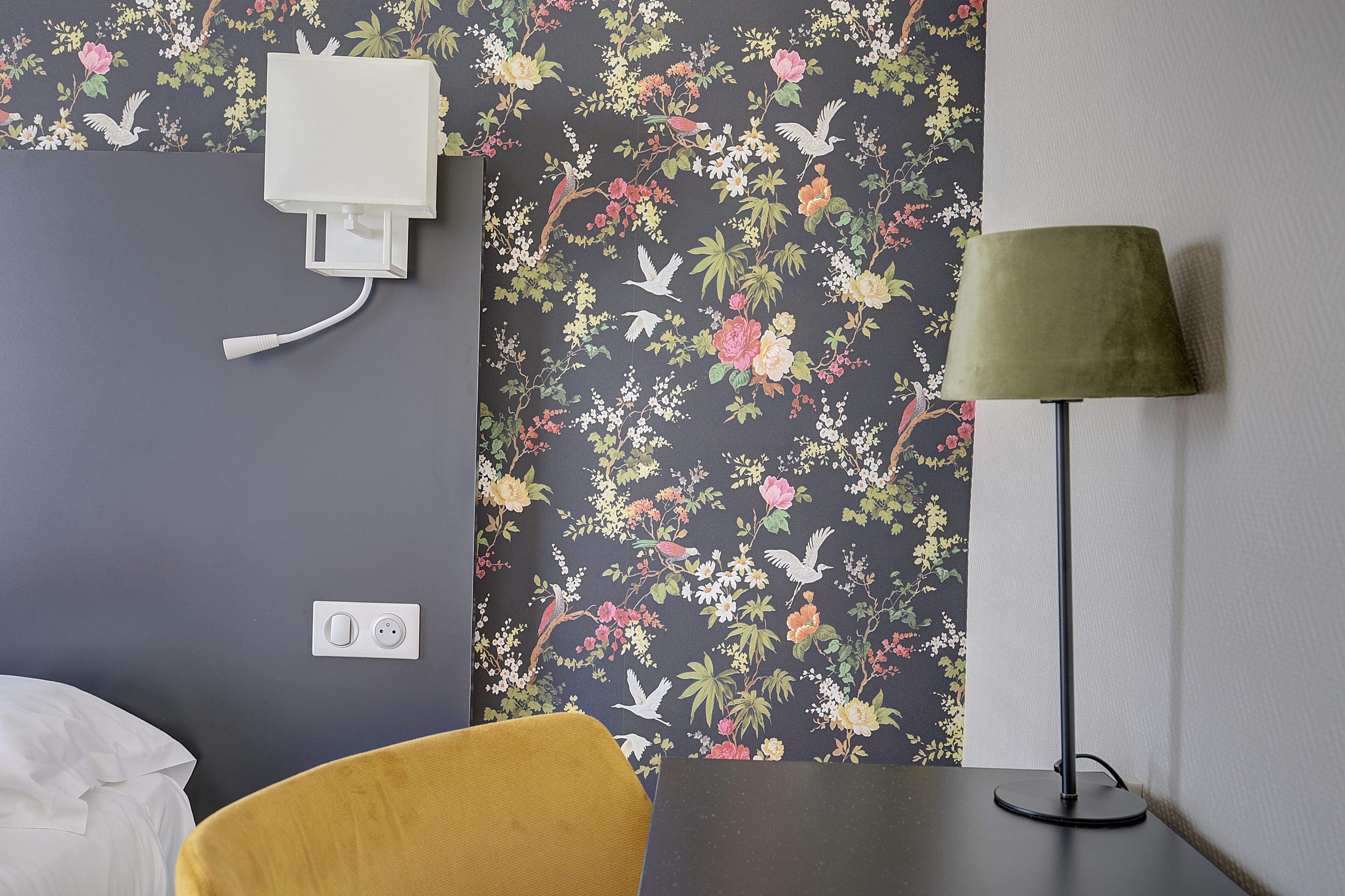 Ein Zimmer mit floralem Wandpapier, einer weißen Steckdose, einem gelben Sessel und einer Stehlampe.