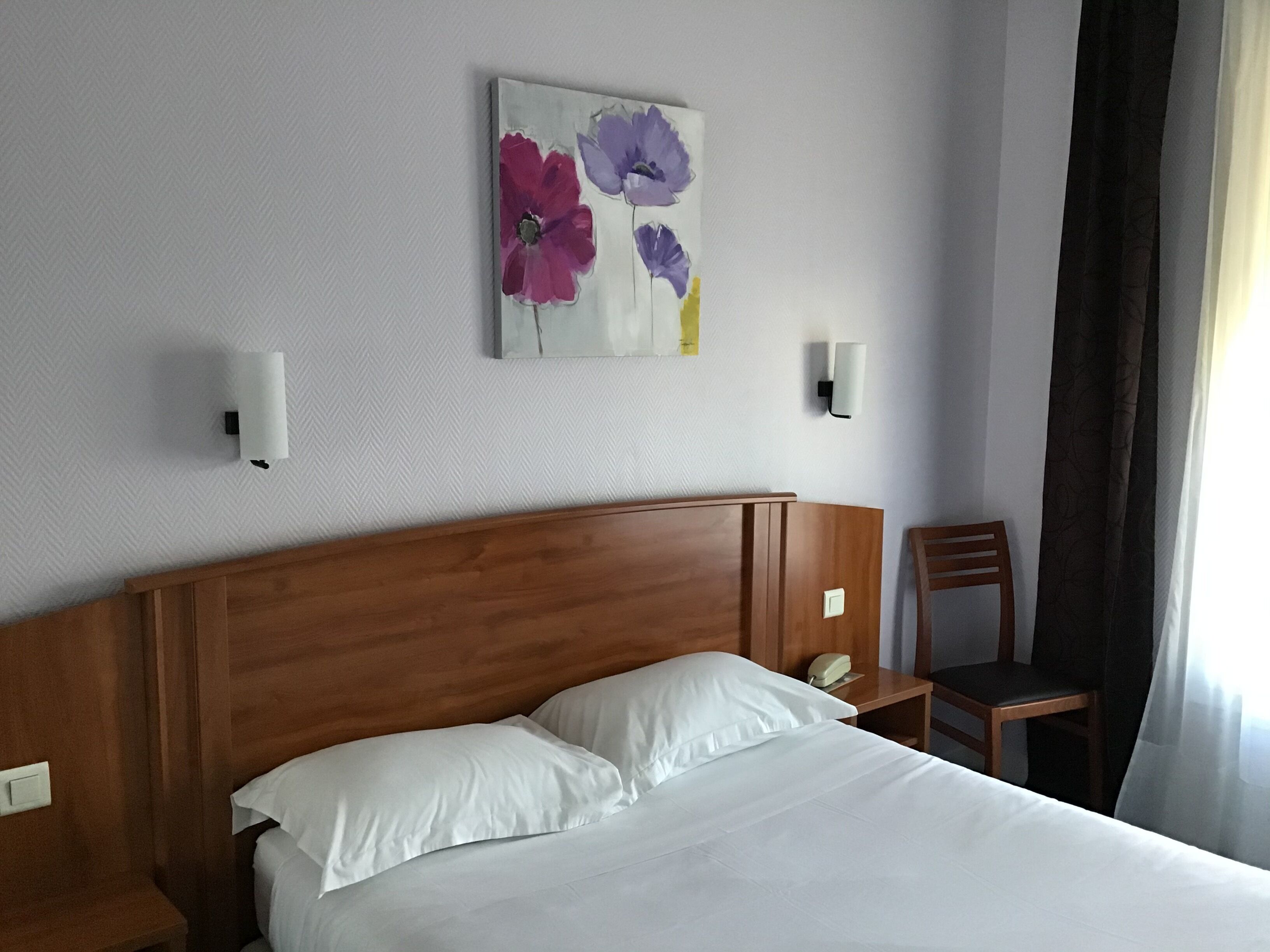 Ein Hotelzimmer mit einem hölzernen Kopfteil, einem Bett mit weißer Bettwäsche, einem Nachttisch mit Telefon und einem an der Wand hängenden Blumenbild.