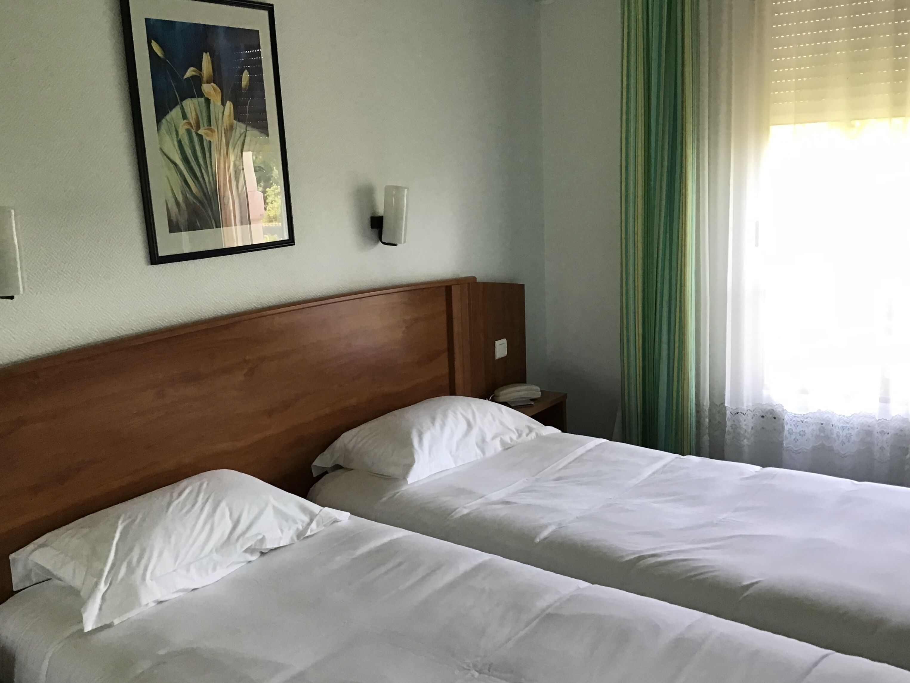 A hotel room with two beds, a wooden headboard, a nightstand, a framed picture on the wall, and a window with curtains.