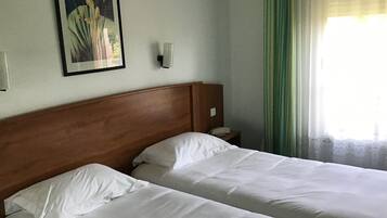 A hotel room with two beds, a wooden headboard, a nightstand, a framed picture on the wall, and a window with curtains.