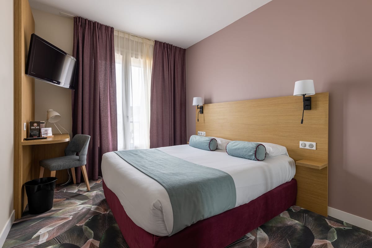 Ein Hotelzimmer mit Bett, Schreibtisch, Stuhl, Fernseher und einem Fenster mit Vorhängen.