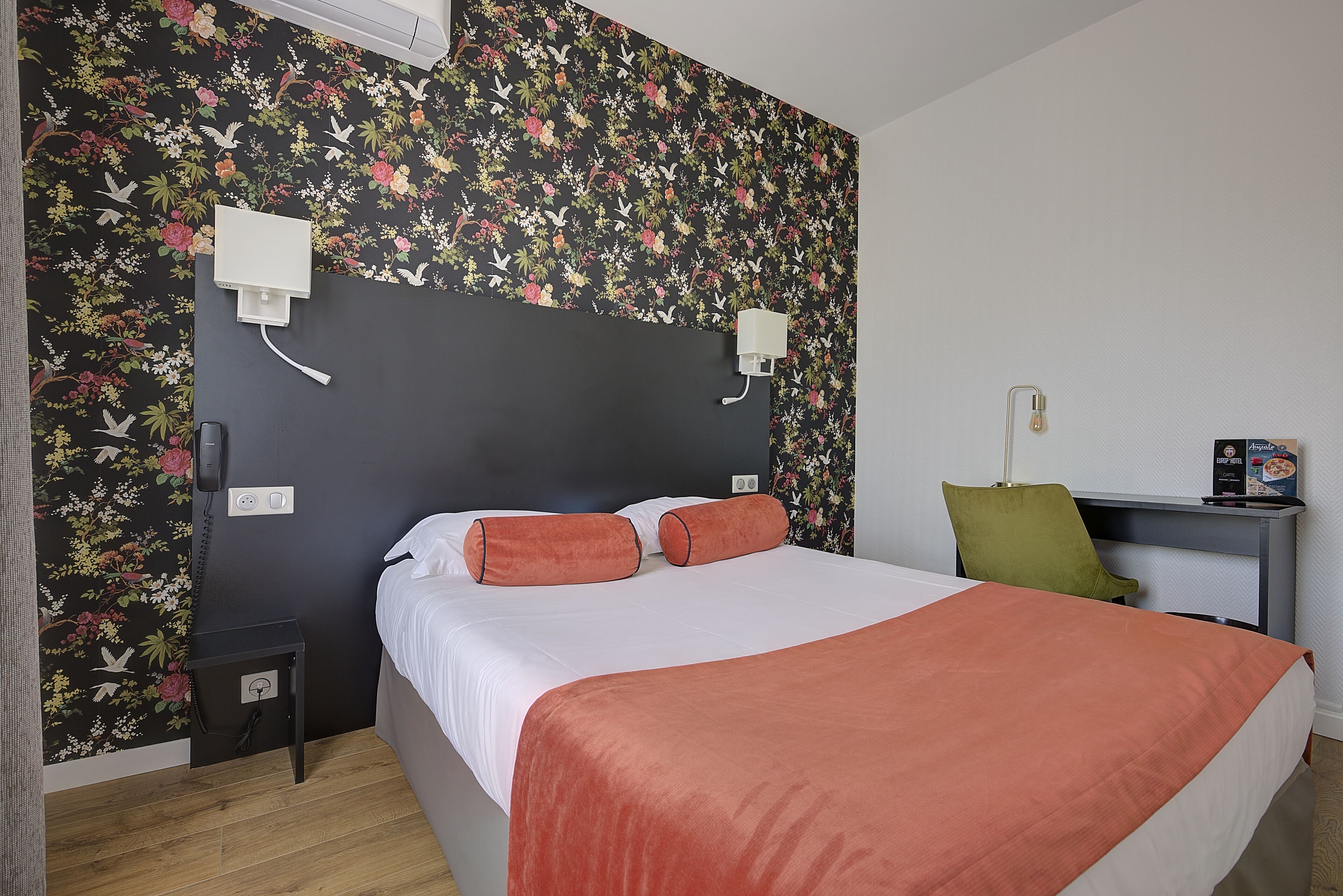 A hotel room with a bed, a desk, a chair, and a wall with floral wallpaper.