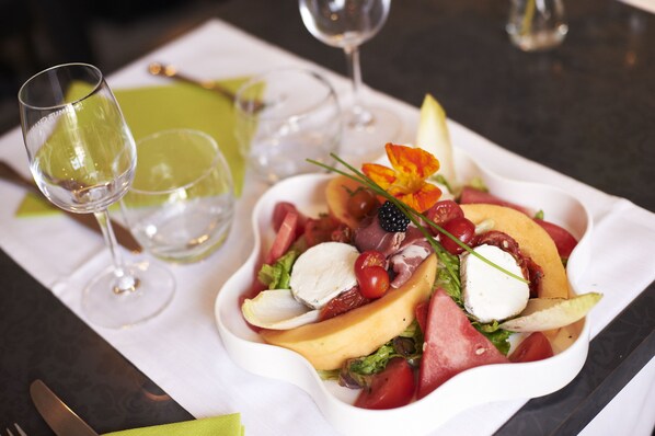 A white plate with a salad containing tomatoes, cheese, and greens, accompanied by two empty wine glasses on a table. - Logis Hotel Restaurant Cristal (Saumur)