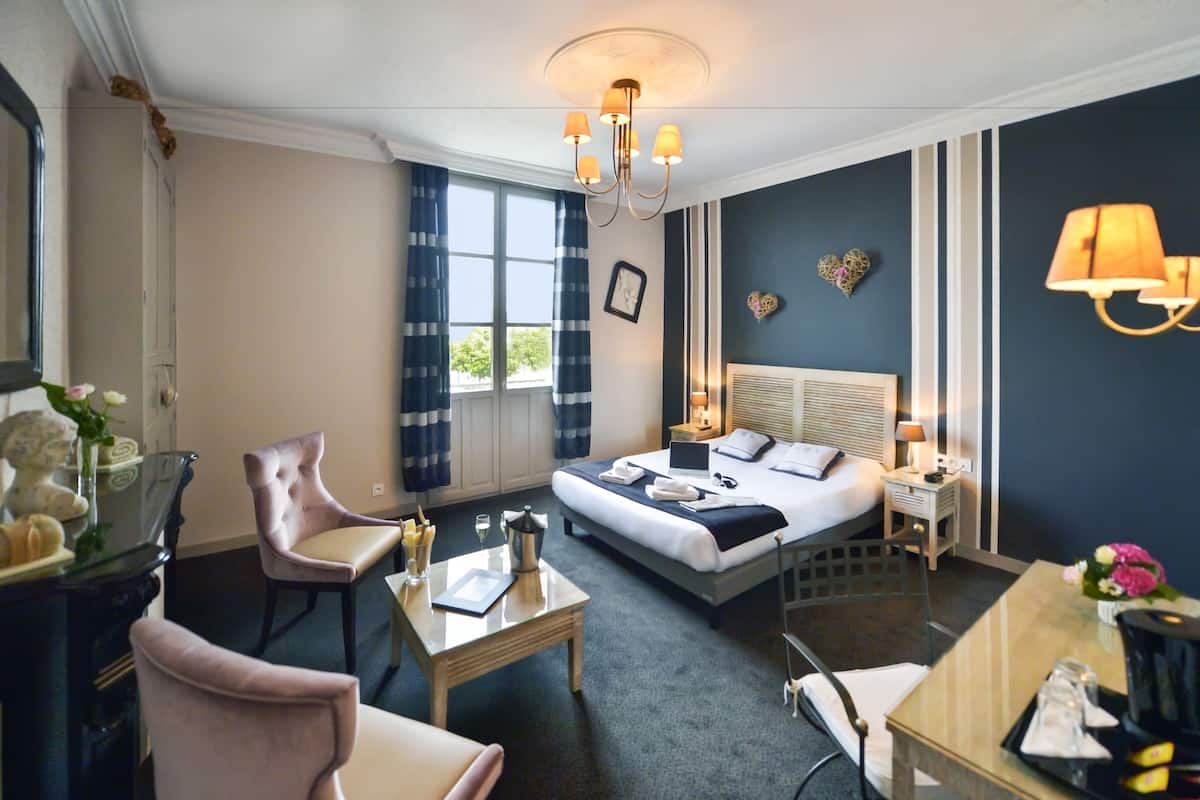 A hotel room with a bed, a desk, a chair, a table, a lamp, and a window with curtains.