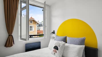 Ein Schlafzimmer mit Bett, einem fensterbepflanzten Fenster, Blick auf Gebäude und einer gelben Wandgestaltung.